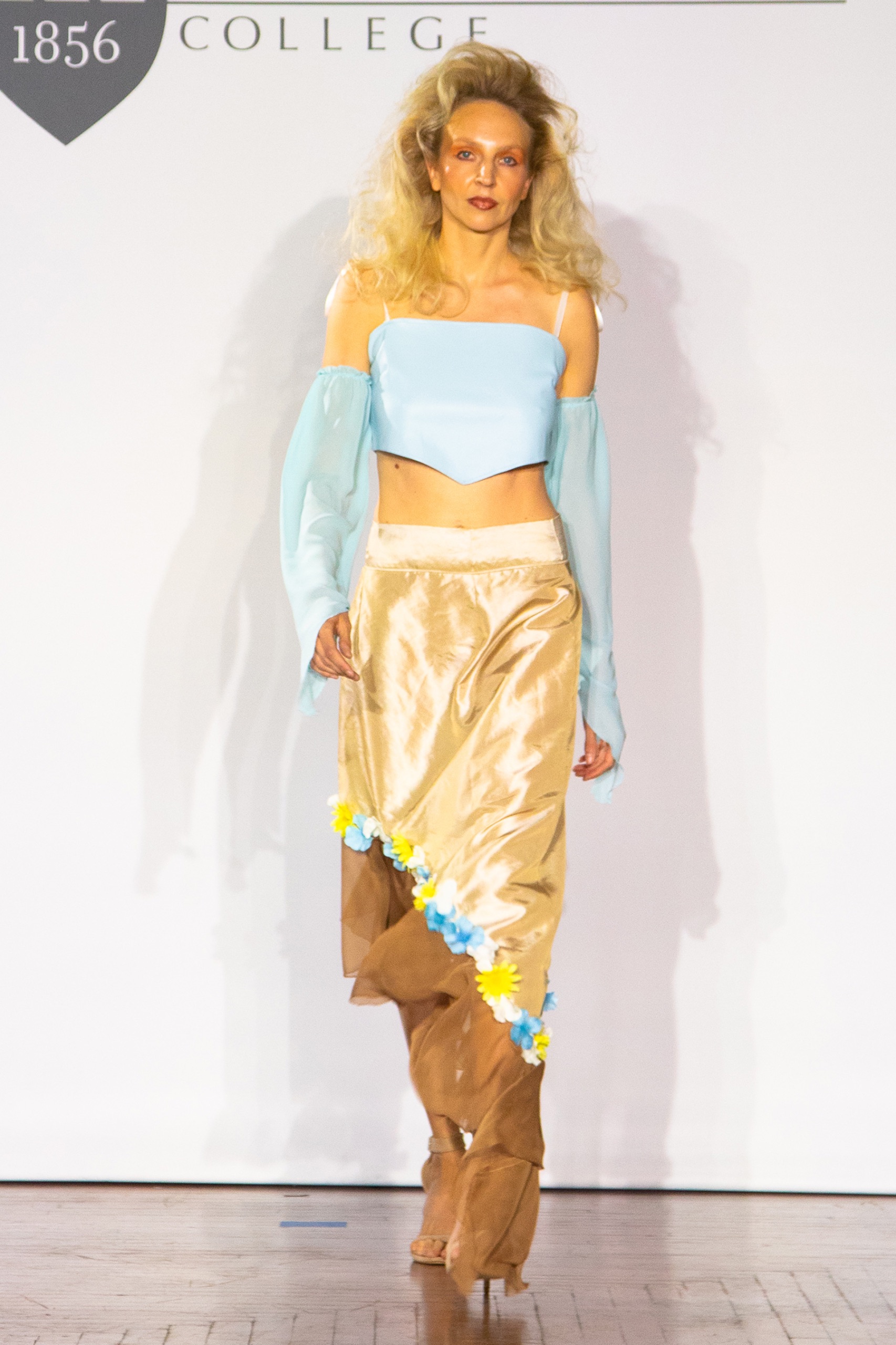 A model walking the runway wearing a gold skirt with a blue top.