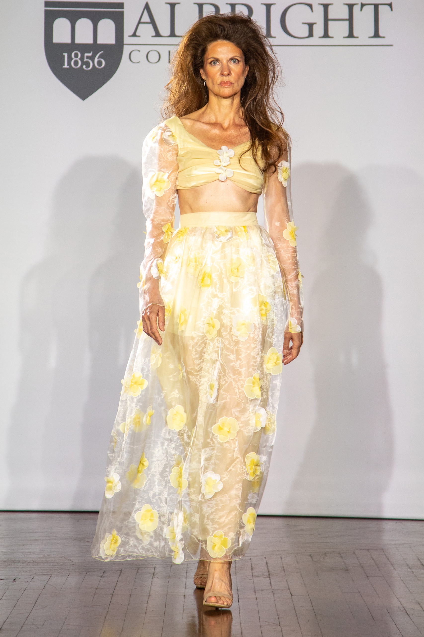 A model walking the runway wearing a yellow top and skirt with a sheer floral overlay.