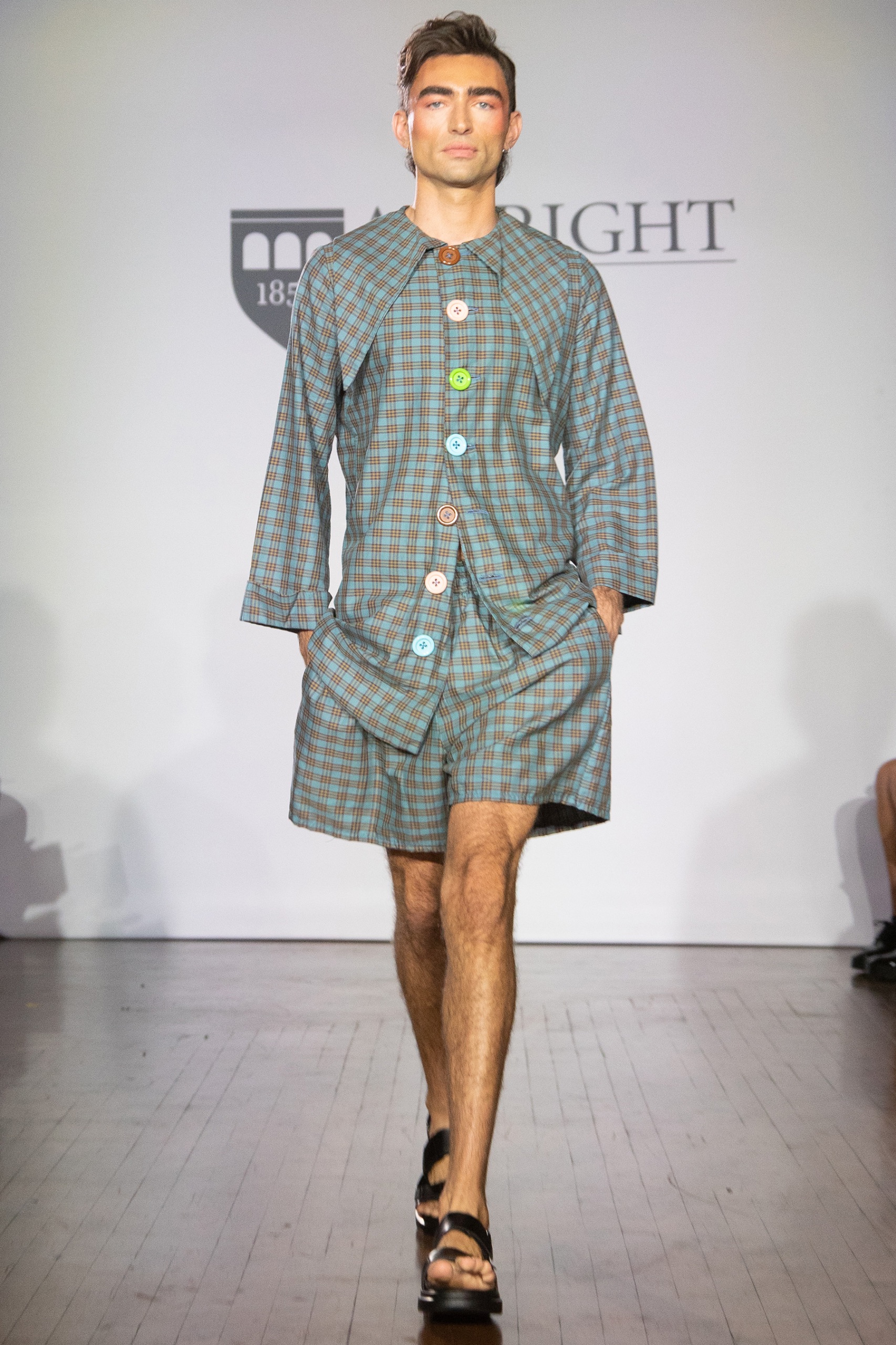 A model walking the runway wearing a long-sleeved checkered dress with buttons.