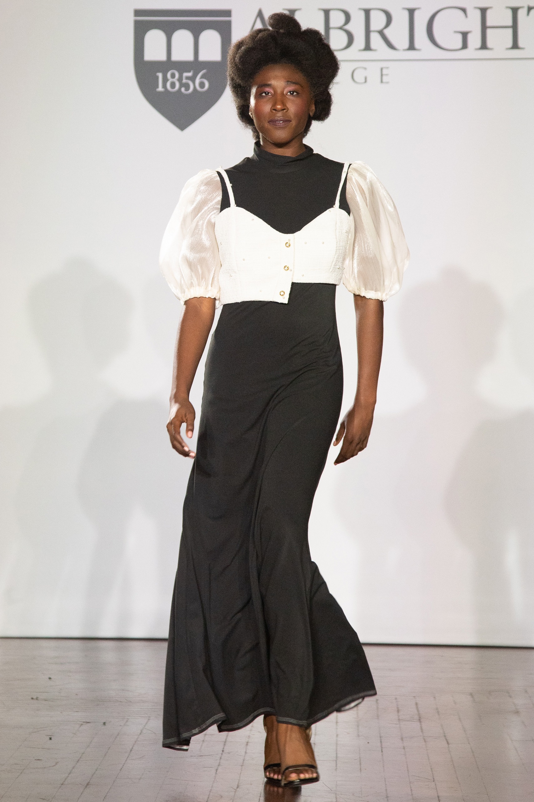 A model walking the runway wearing a long black dress with white puffy sleeves and a white vest.