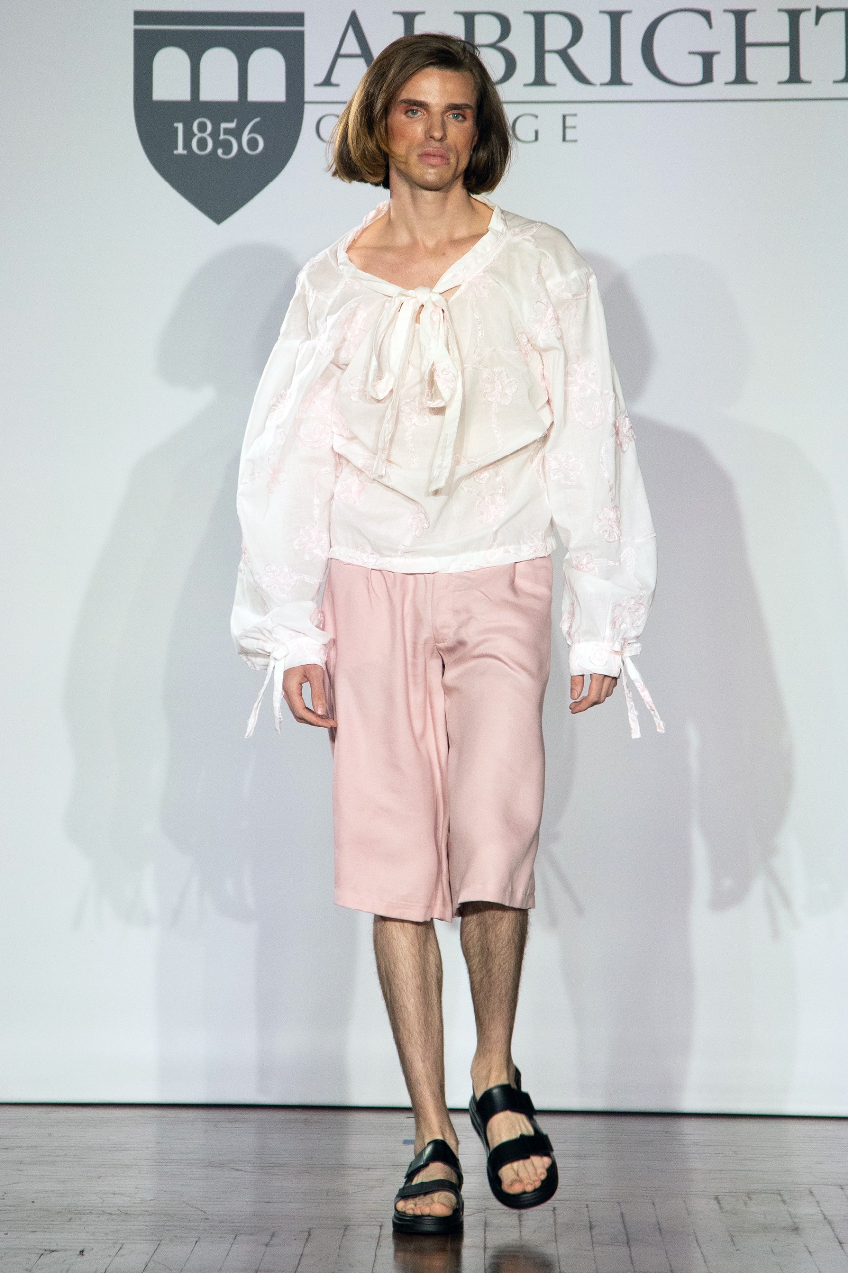 A model walking the runway wearing a long-sleeved white skirt, pink shorts and black shoes.