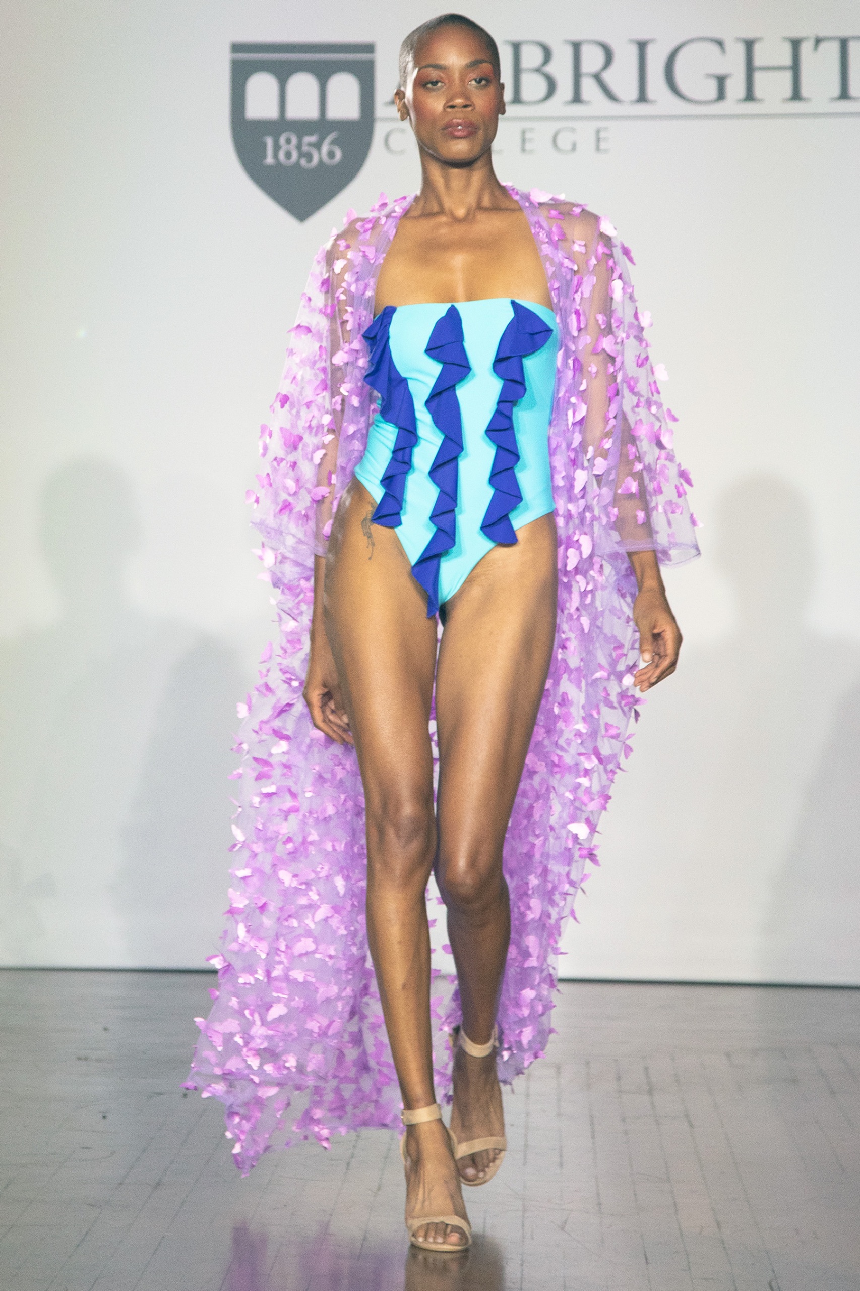 A model walking the runway wearing a blue striped bathing suit with long purple robe.