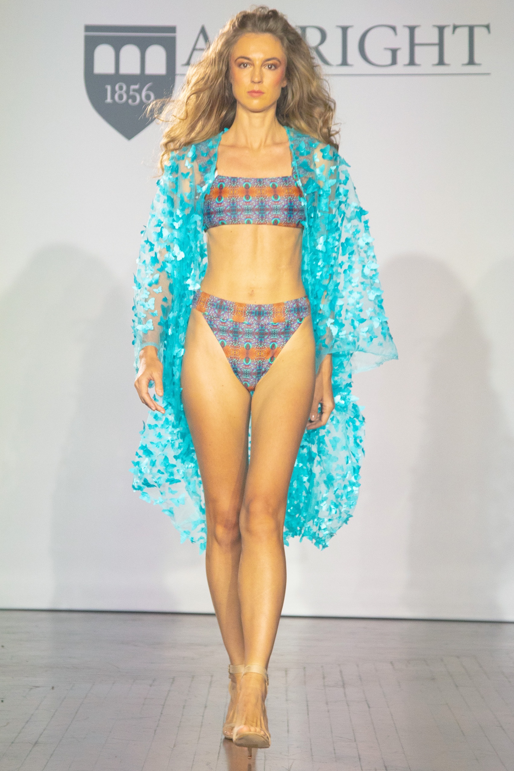 A model walking the runway wearing a blue and orange patterned bikini with long blue robe.