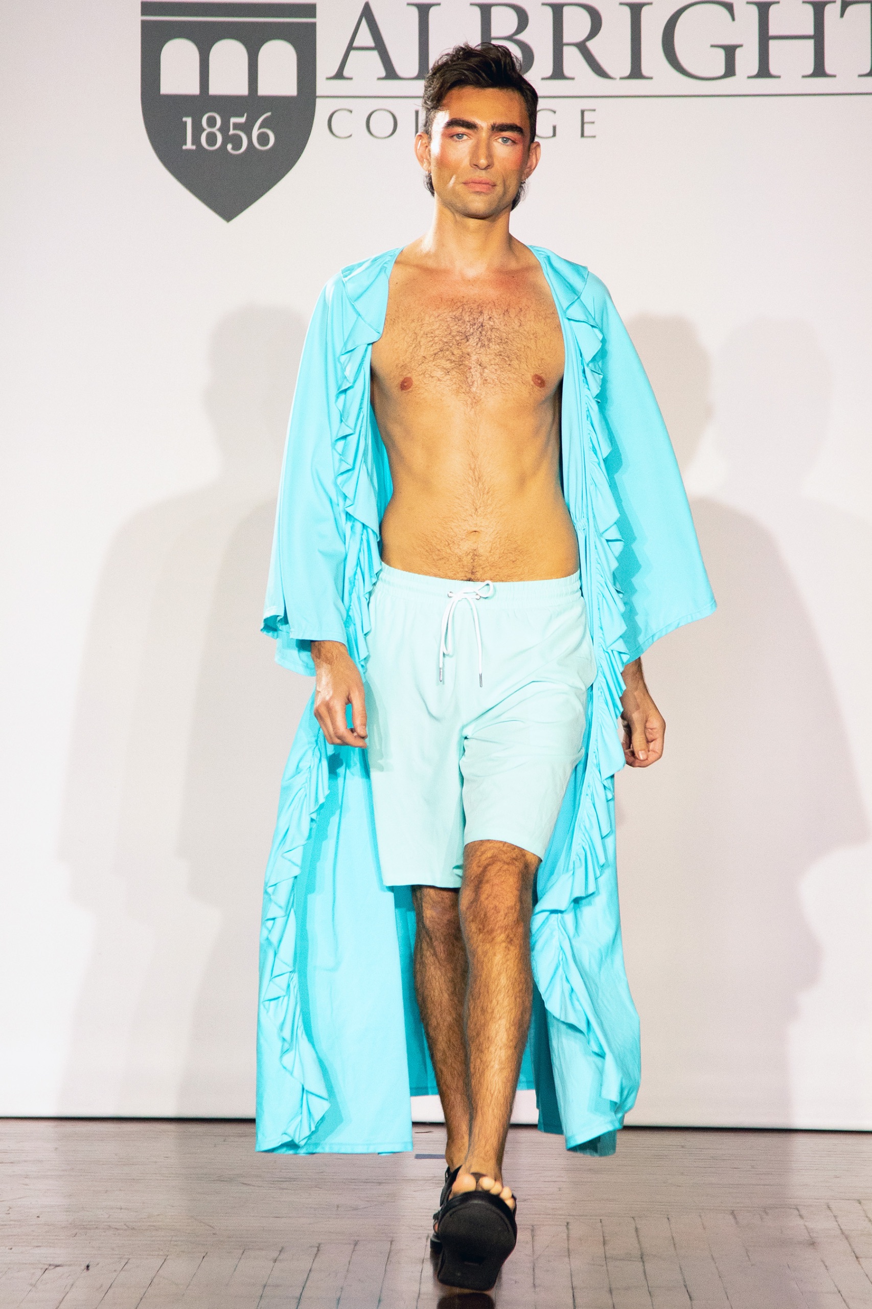 A model walking the runway wearing blue swim shorts and a long blue robe.