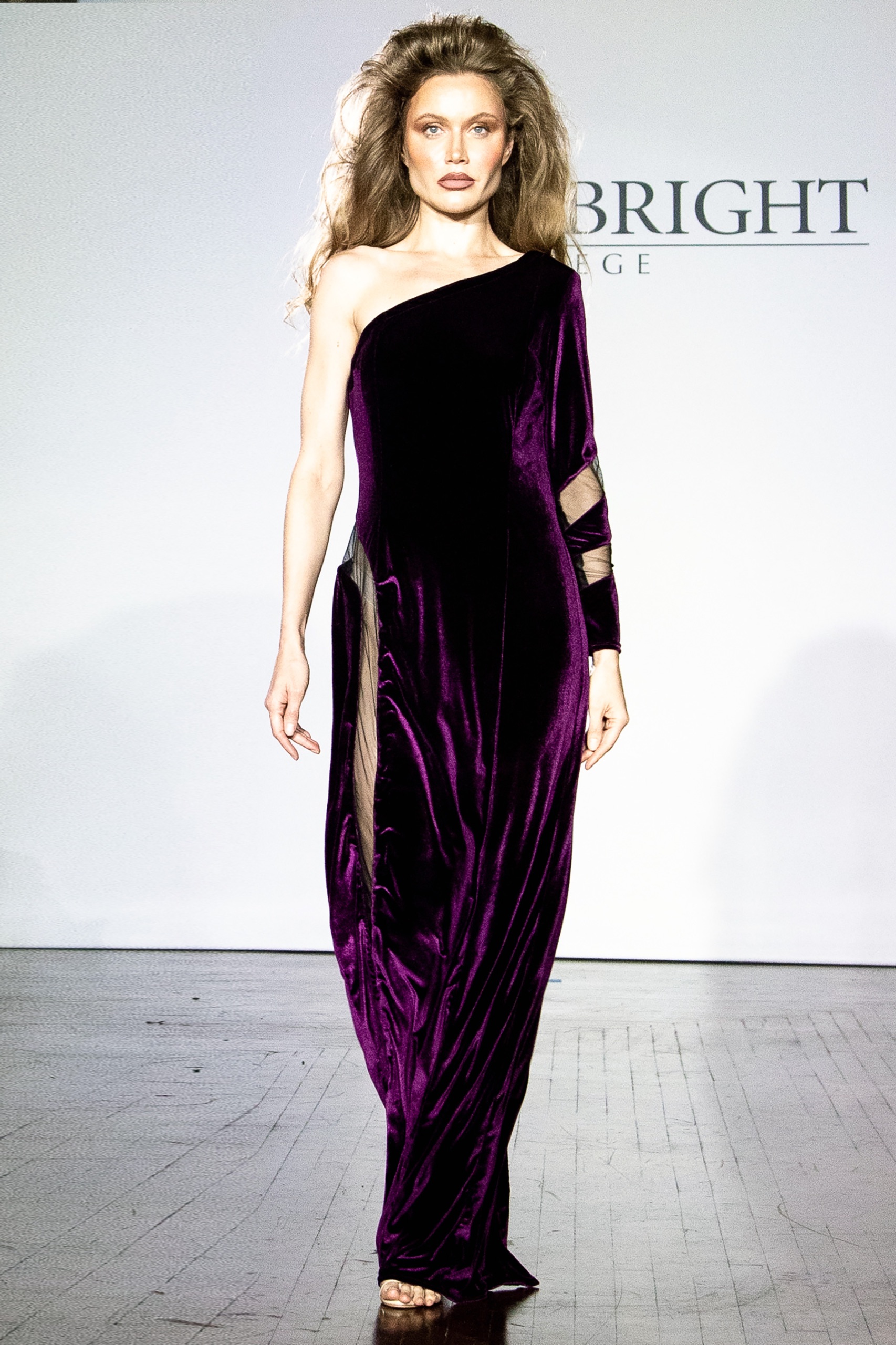 A model walking the runway wearing a long one-shoulder purple dress.