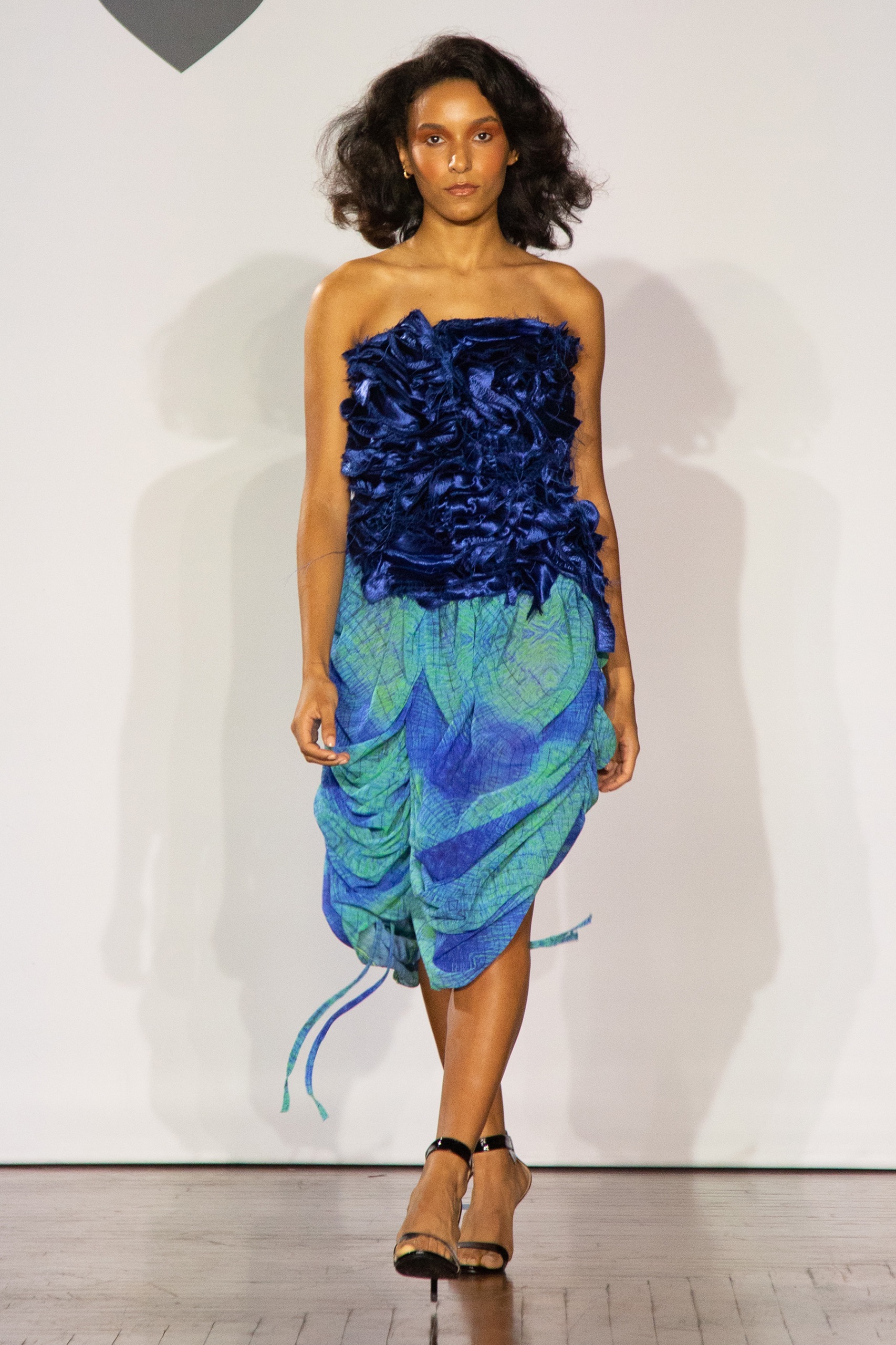 A model walking the runway wearing a dark blue top with a green and blue skirt.