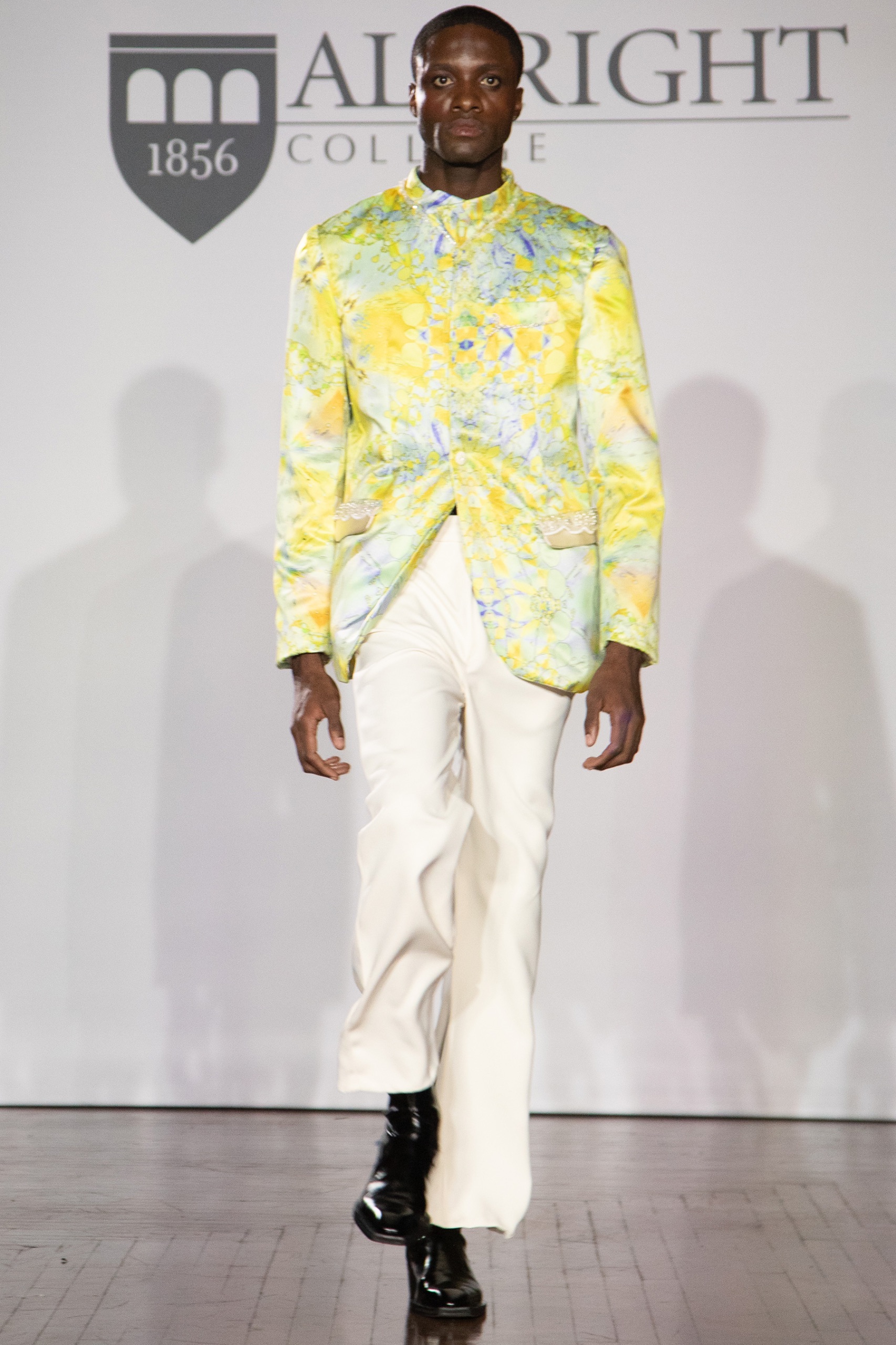 A model walking the runway wearing a yellow and green blazer with white pants.