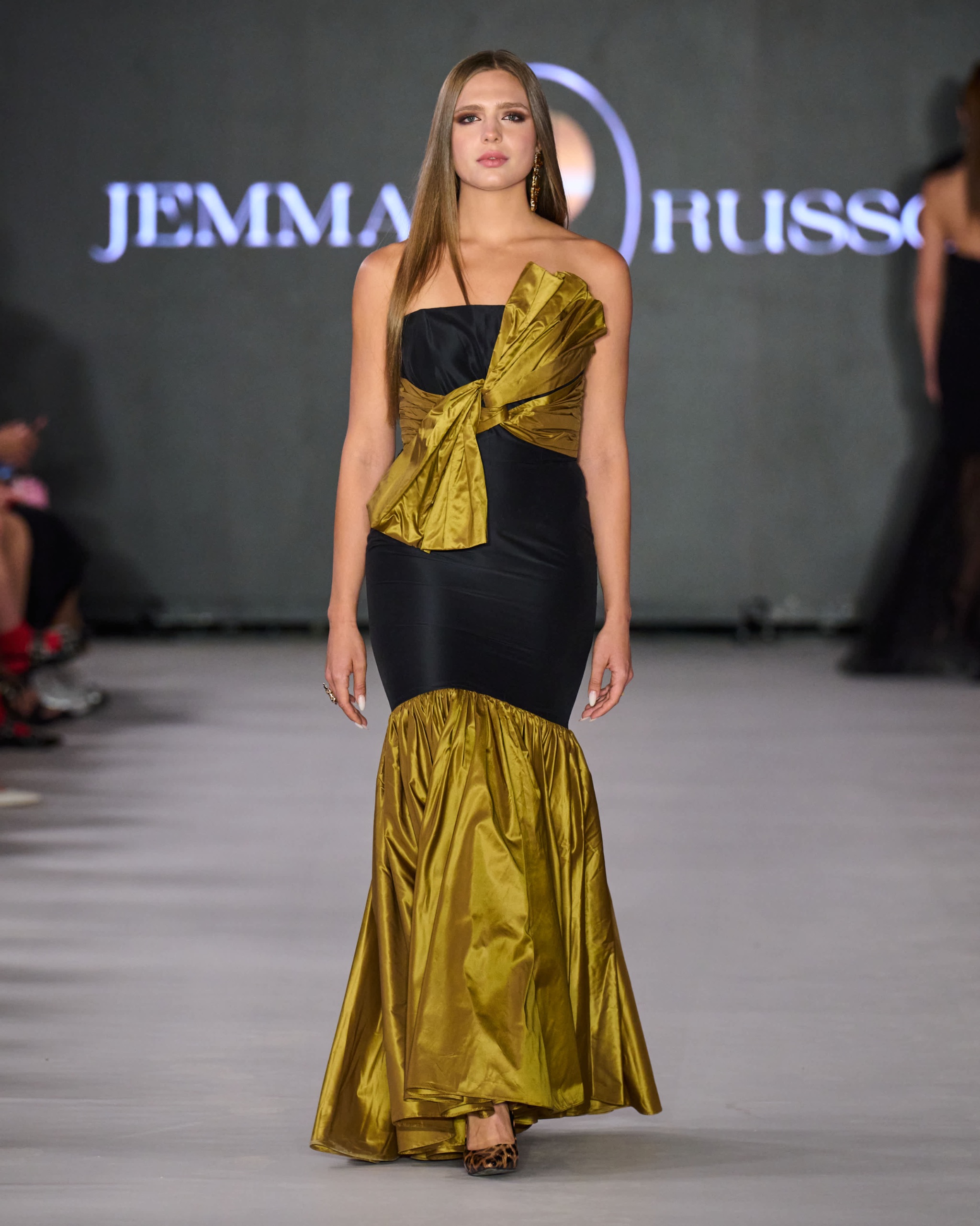 A model walking the runway wearing a long black and gold dress with bow detail.