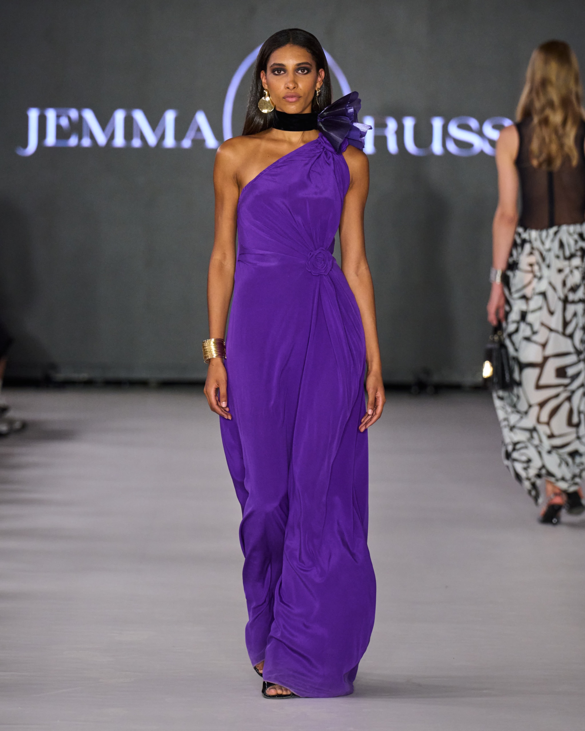 A model walking the runway wearing a long one-shoulder purple dress.