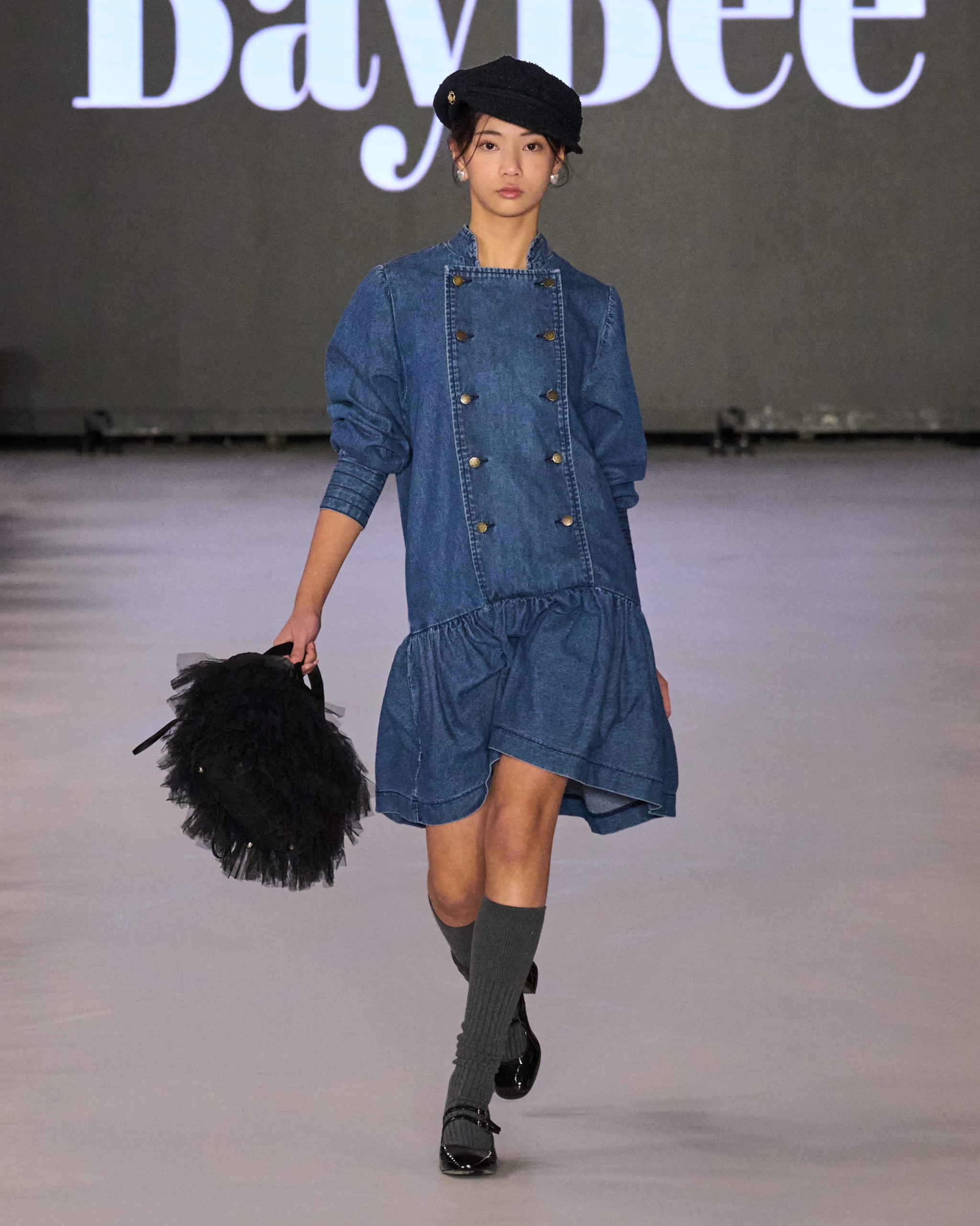 A model walking the runway wearing a blue dress holding black bag.