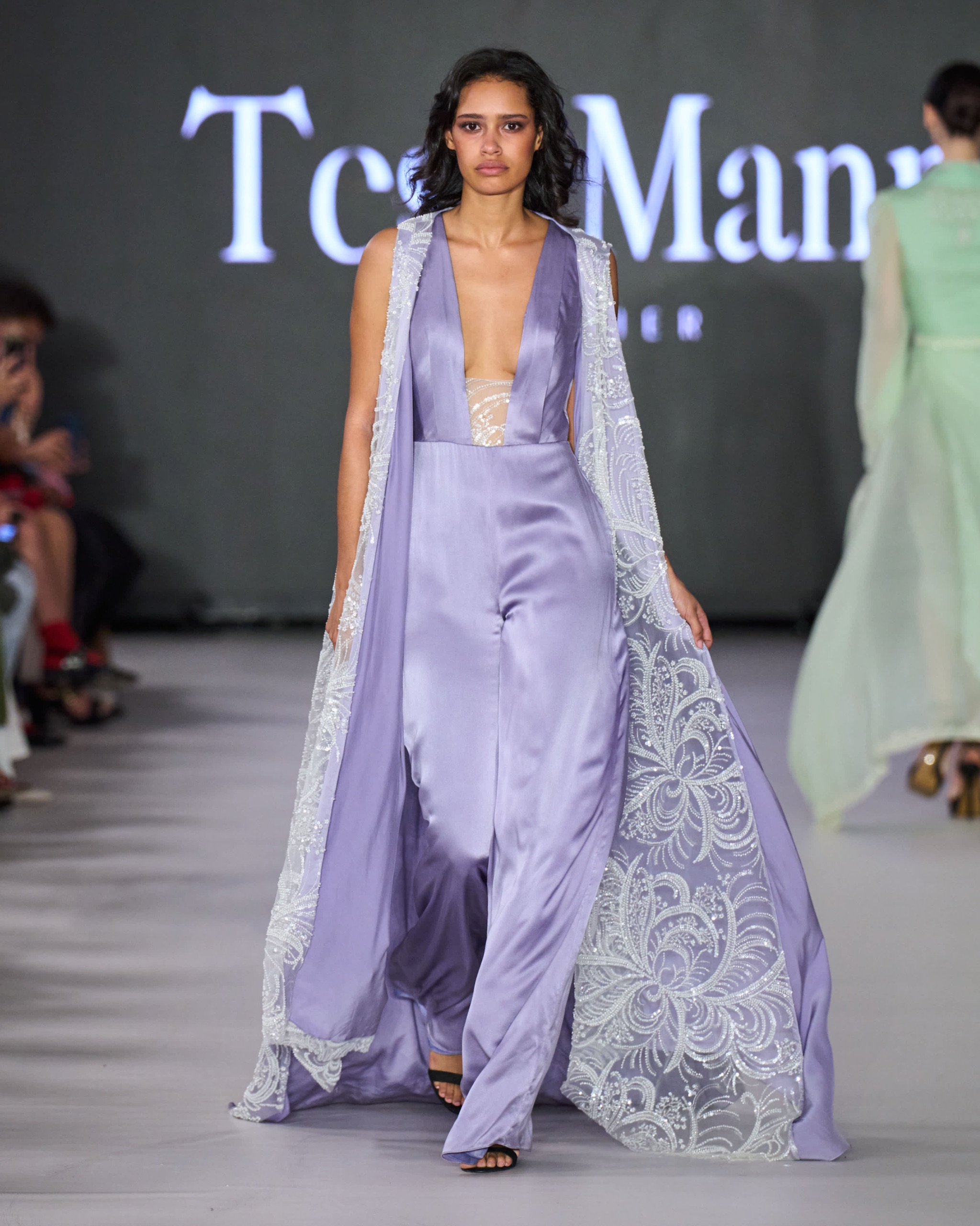 A model walking the runway in a flowy lavender gown with lace detailing.