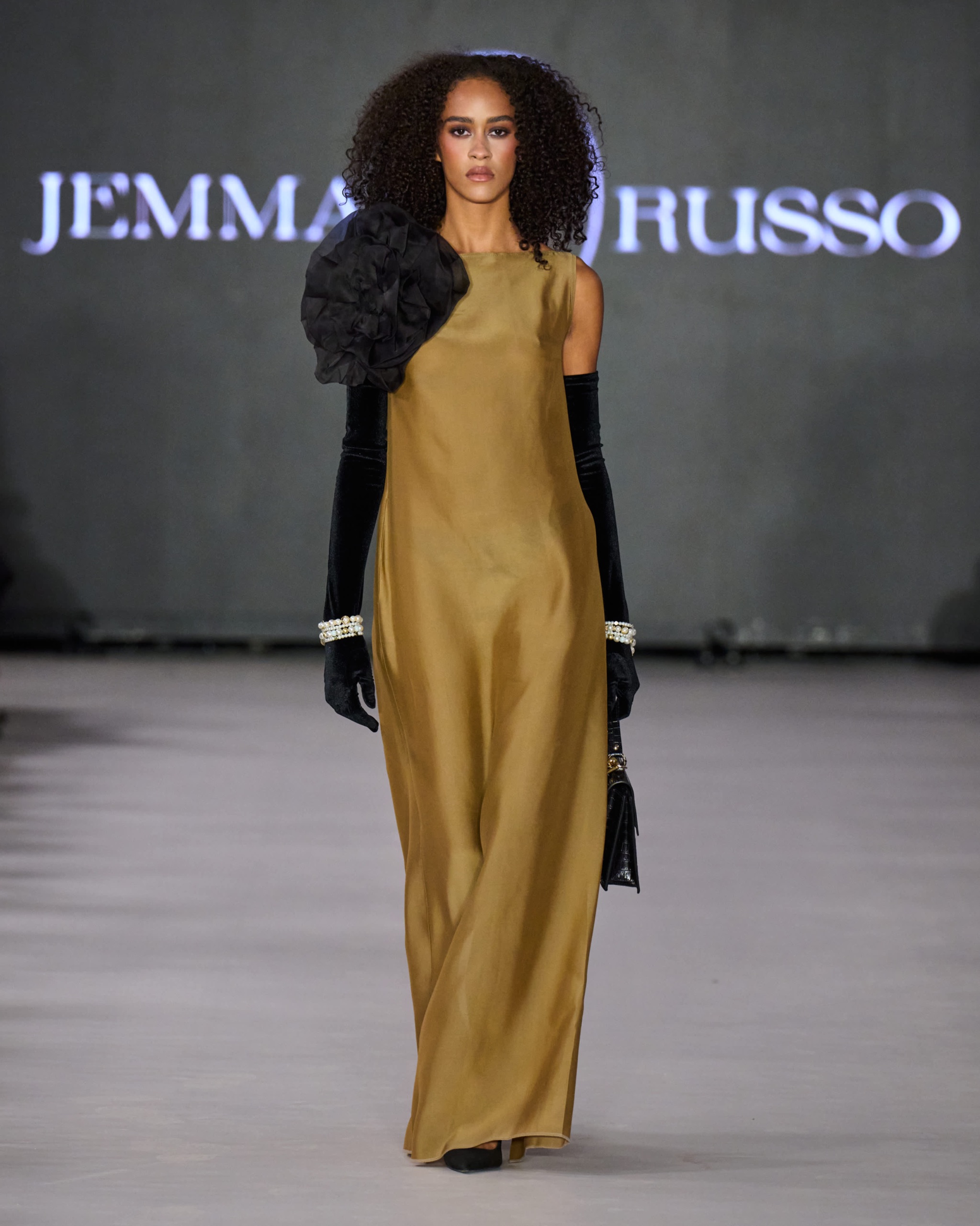 A model walking the runway wearing a long gold dress with black gloves.