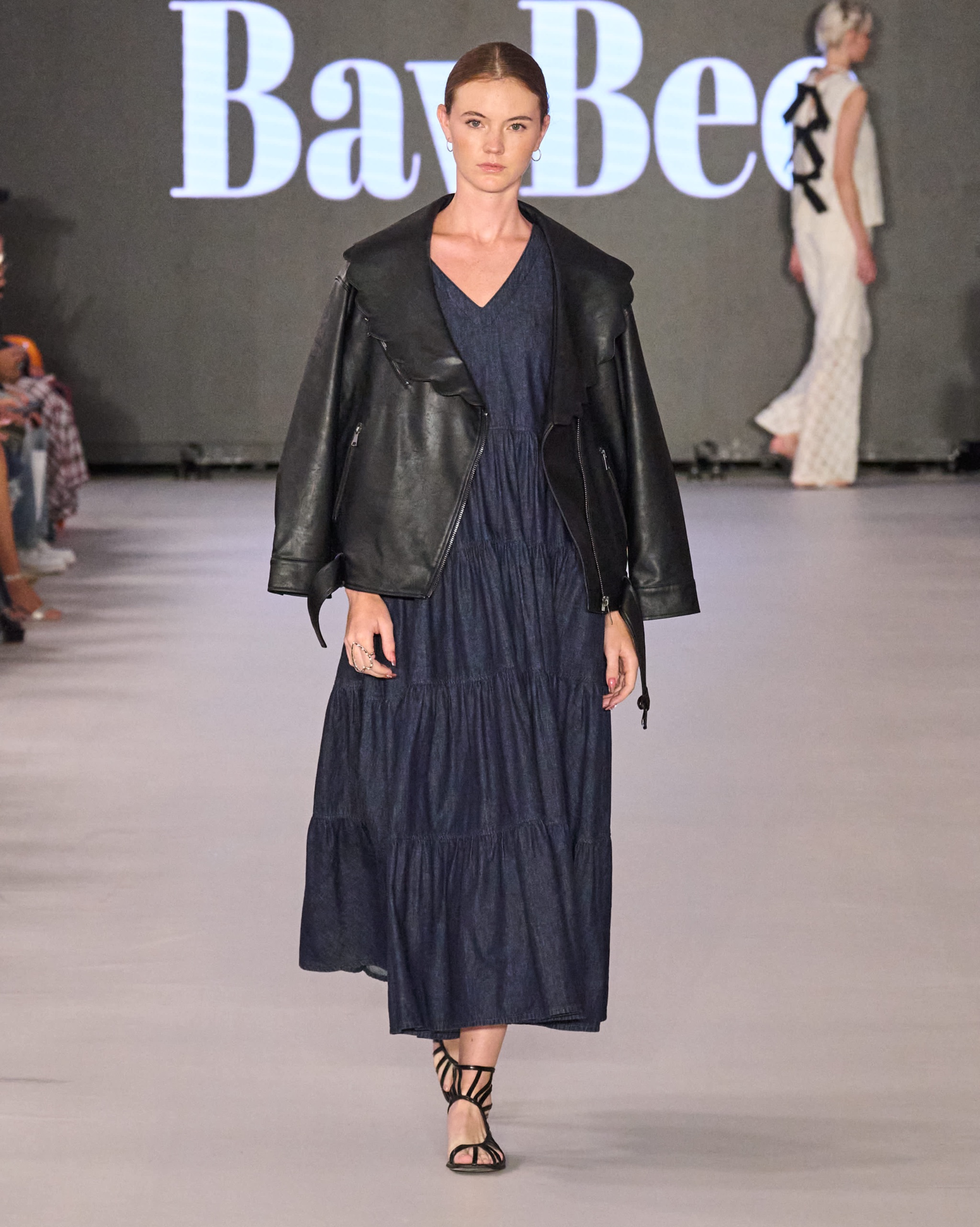 A model walking the runway wearing a long navy dress with a black jacket.