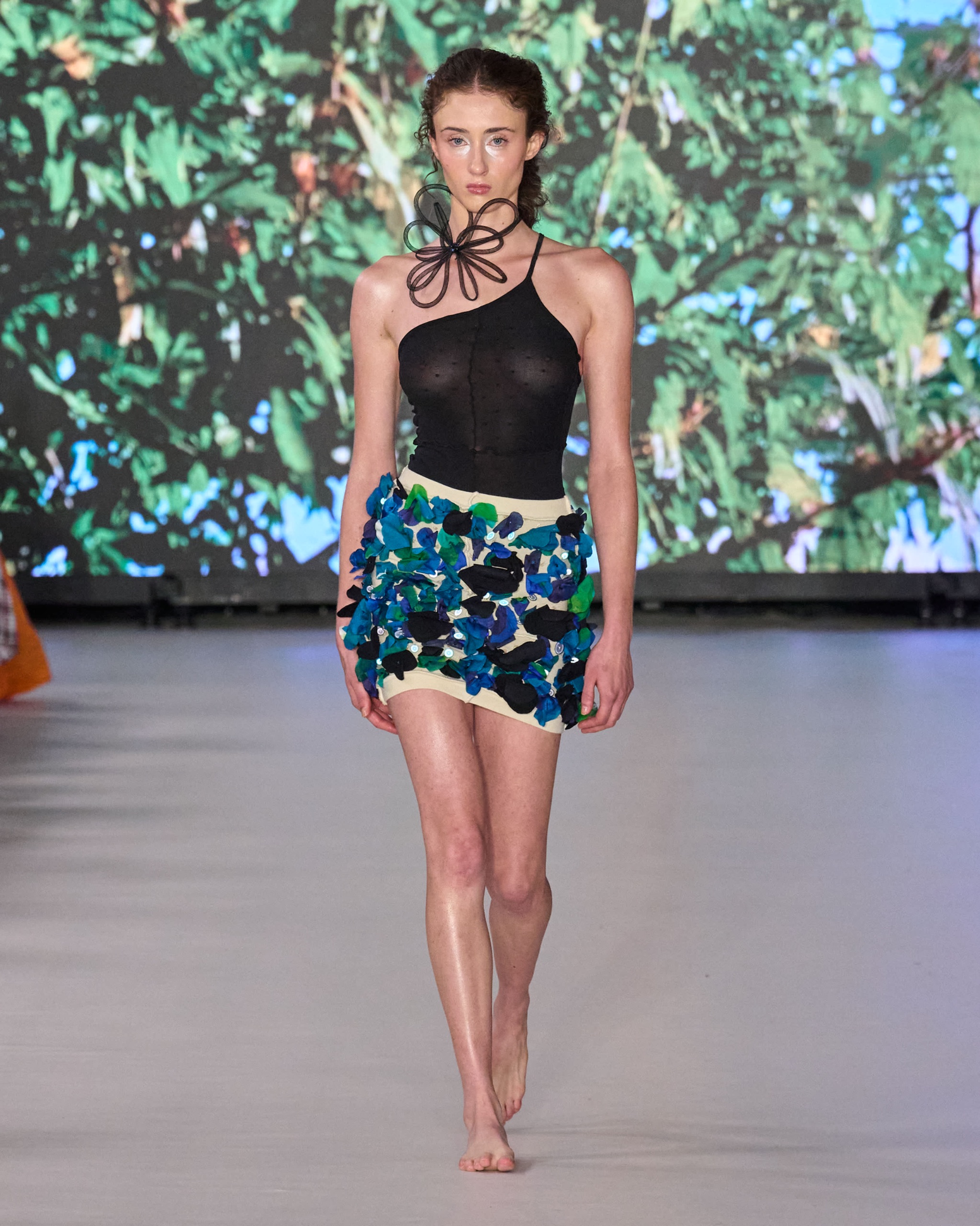 A model walking the runway wearing a black top and blue and green patterned skirt.