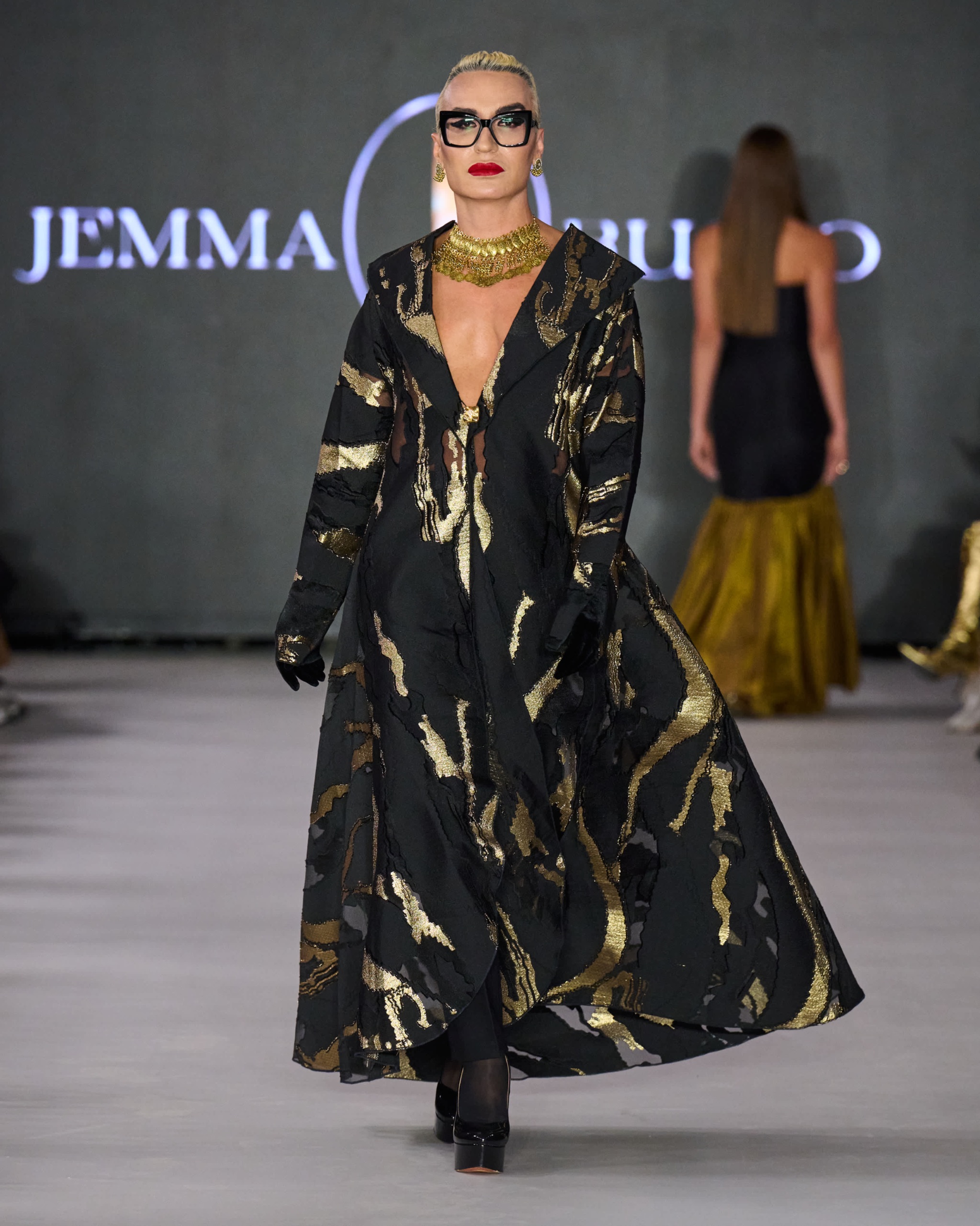 A model walking the runway wearing a long black dress with gold detailing.