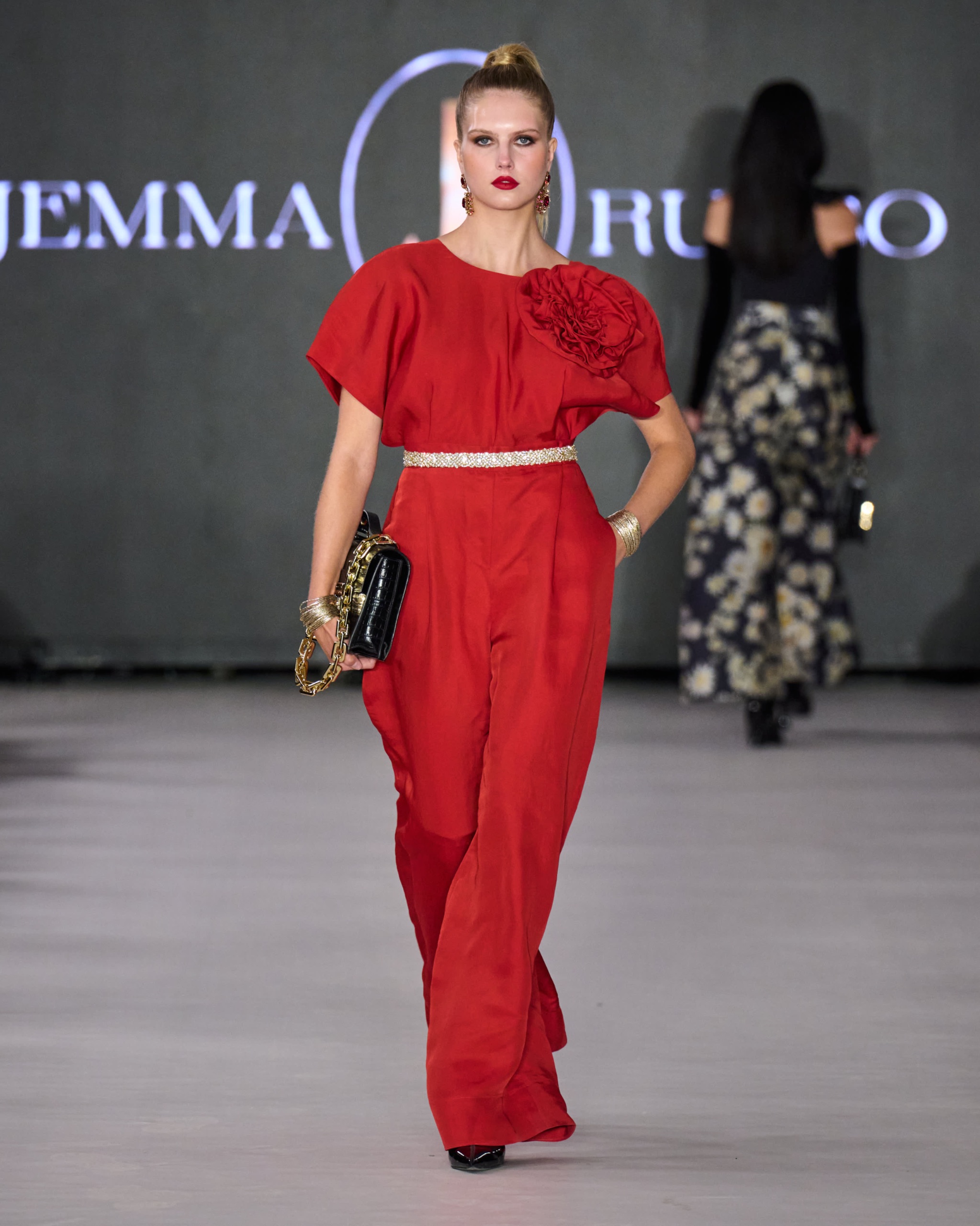 A model walking the runway with a red top and red pants holding a bag.