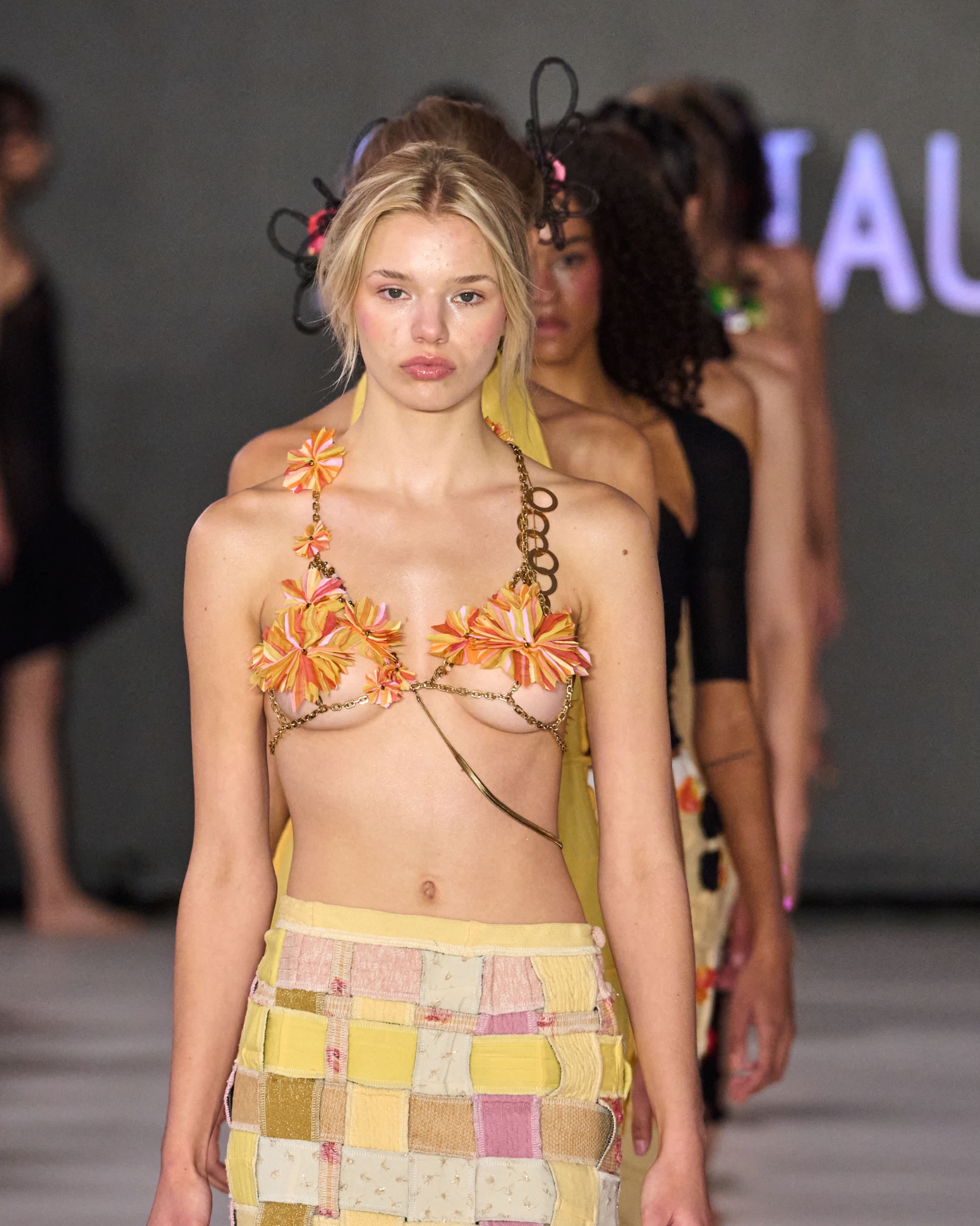 A model walking the runway wearing a orange bikini top with a yellow checkered skirt.