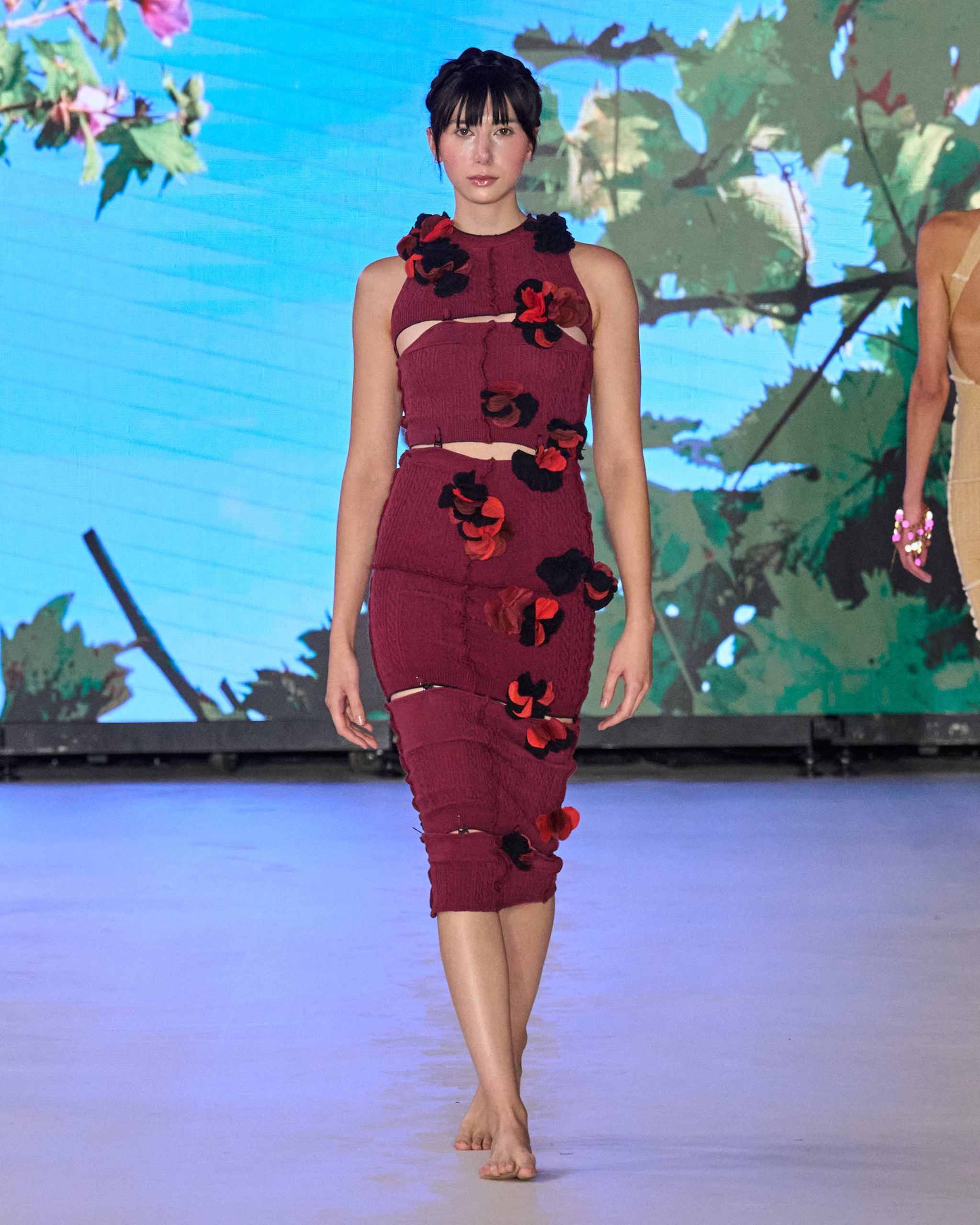 A model walking the runway wearing a burgundy dress with detailed embellishments.