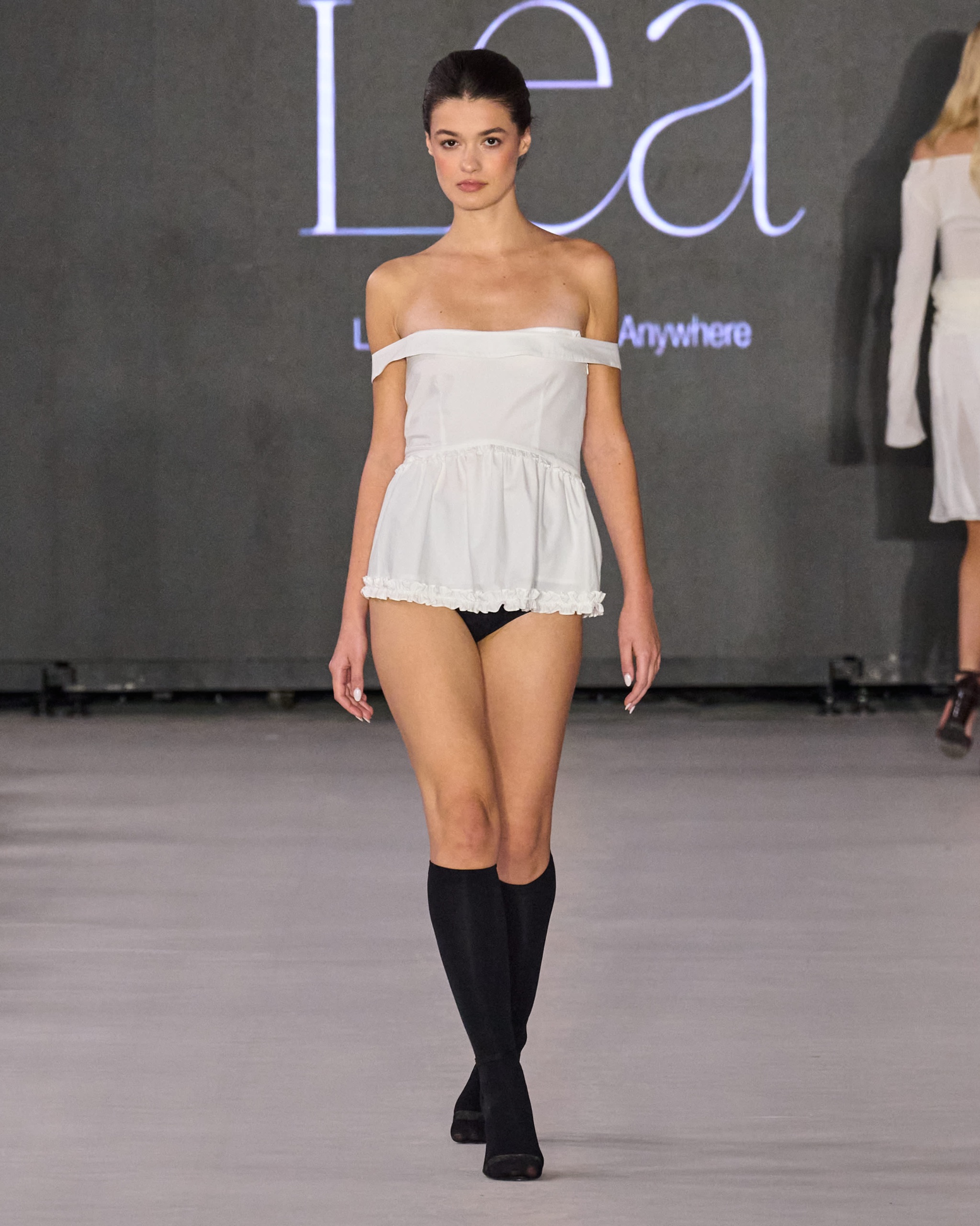 A model walking the runway wearing a strapless white dress with long black boots.