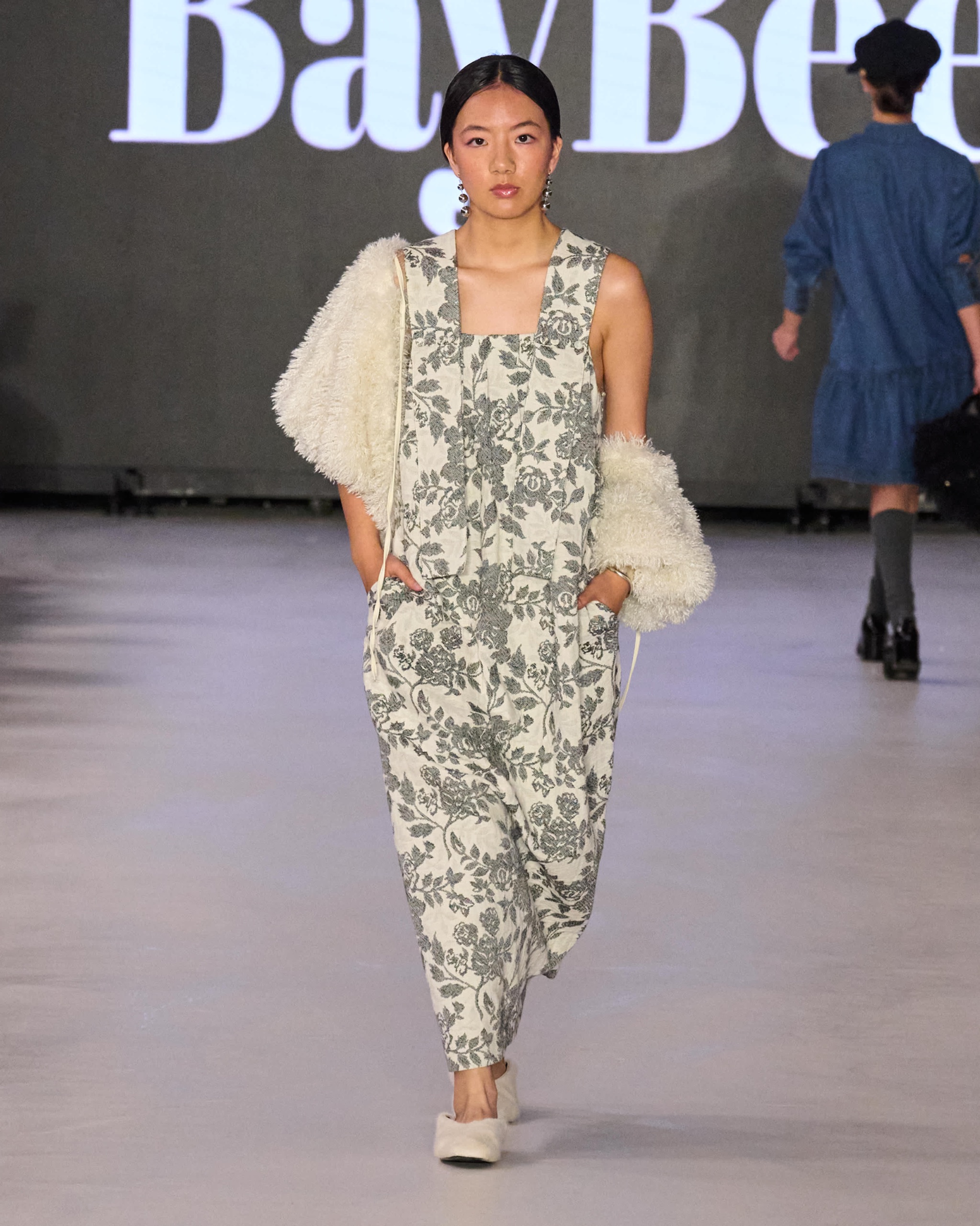 A model walking the runway wearing a floral-patterned jumpsuit with a fur-lined vest.