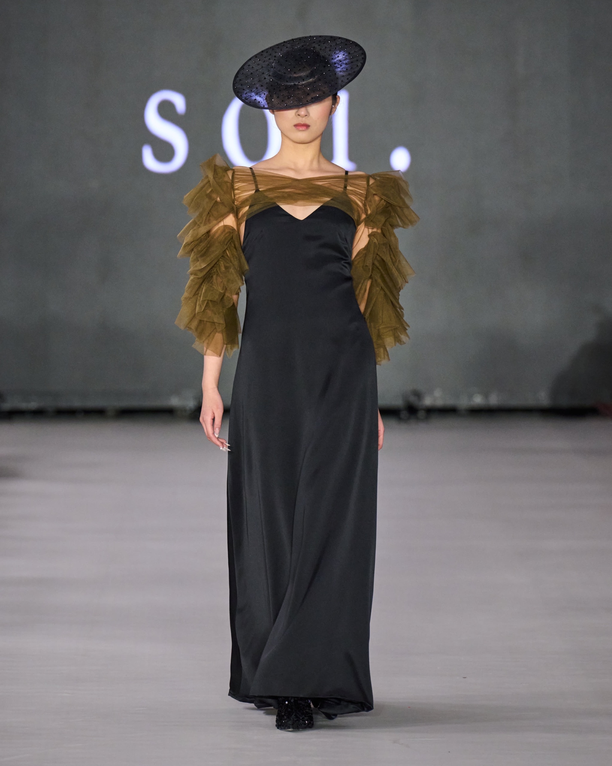 A model walking the runway wearing a long black dress with fur sleeves.