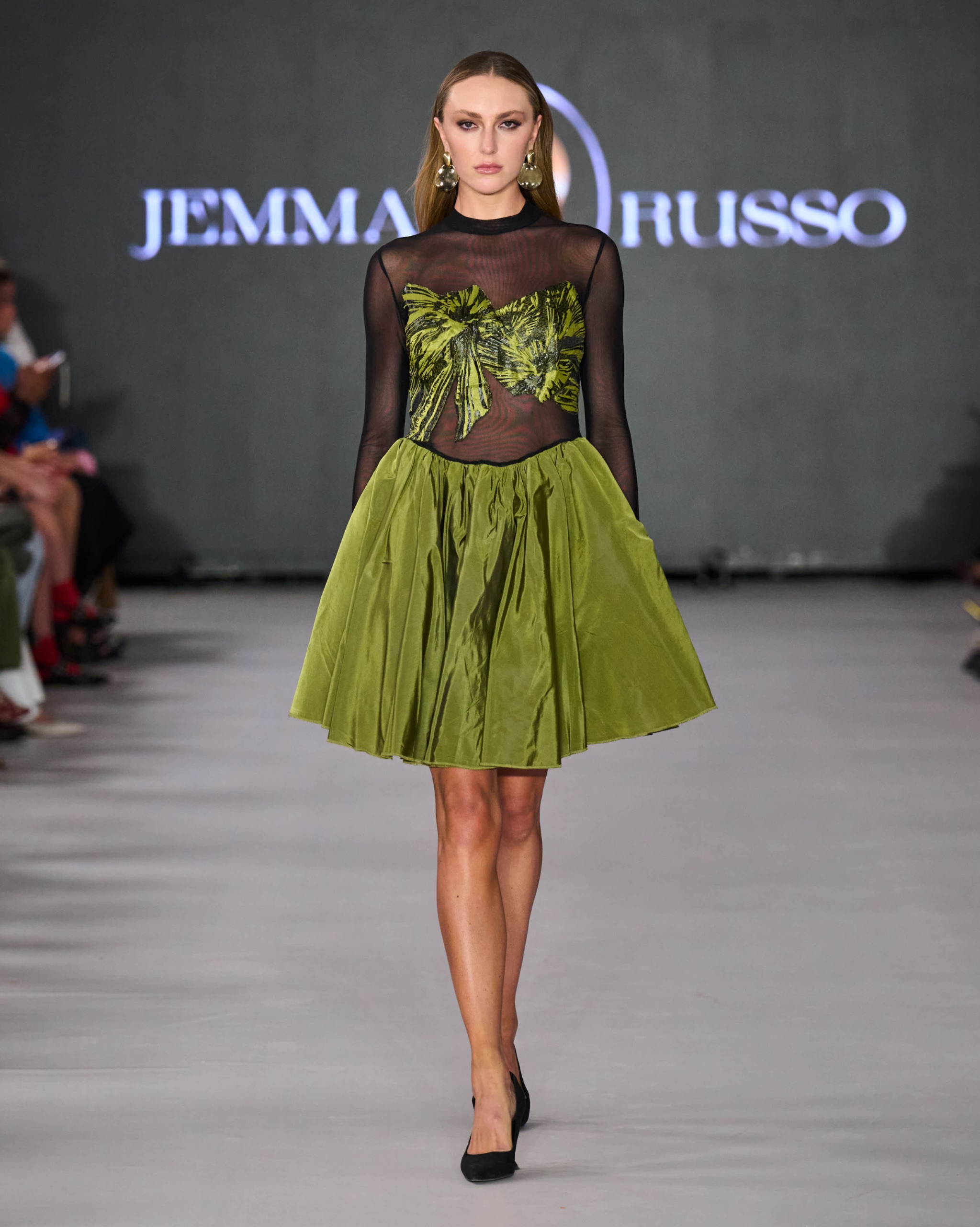 A model walking the runway wearing a green skirt and top with a sheer black top.