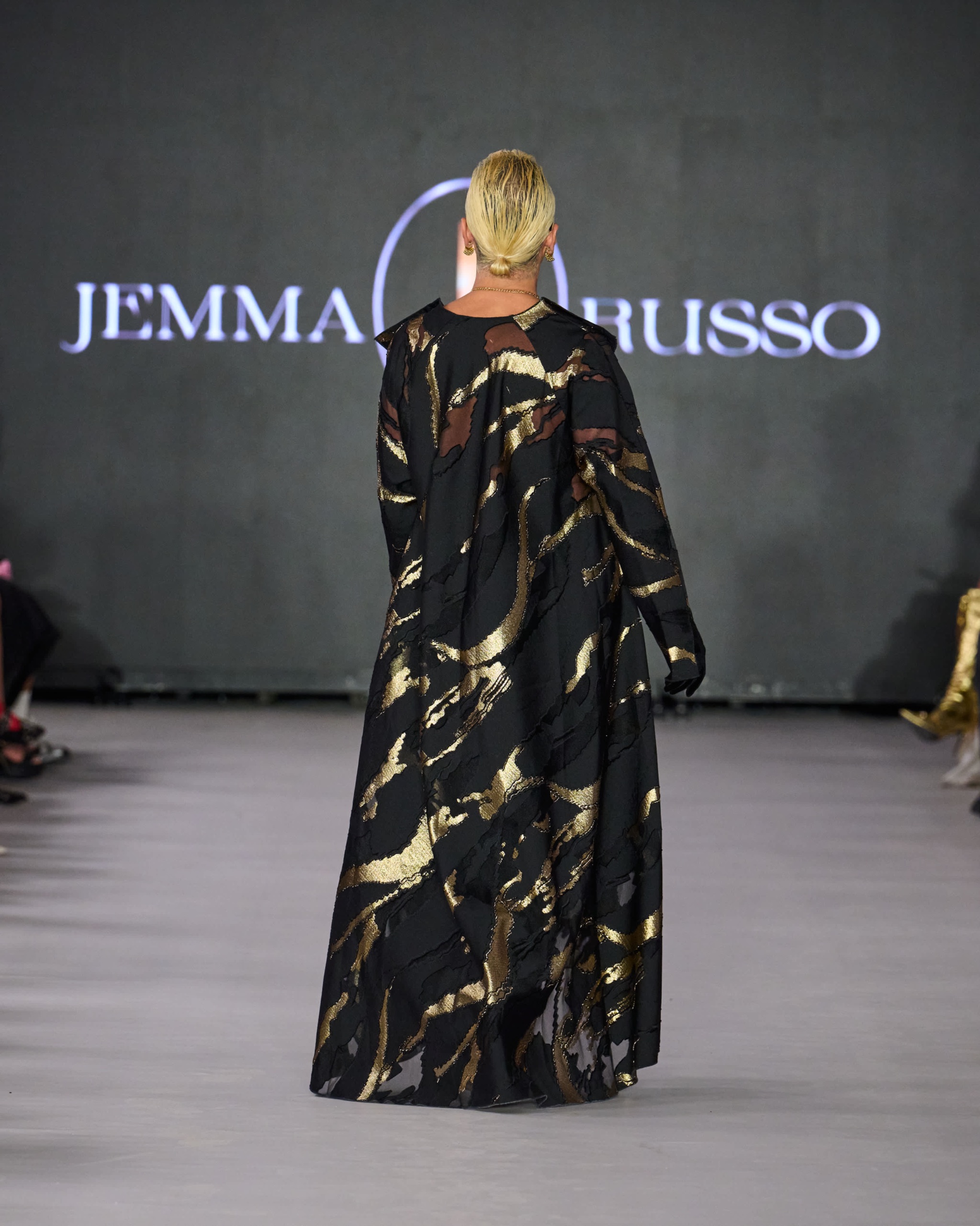 A model walking on the runway wearing a long black dress with gold detailing.