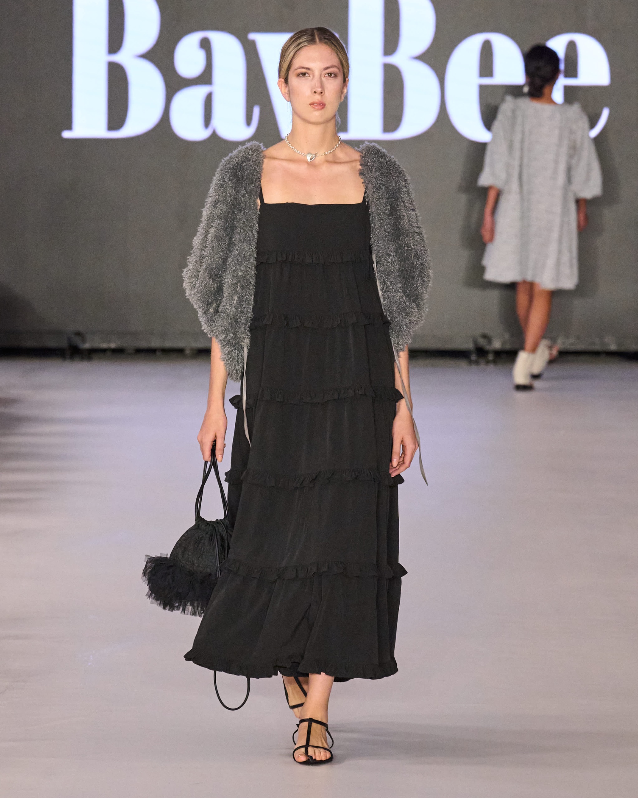 A model walking on the runway wearing a long black dress, grey jacket and black bag.