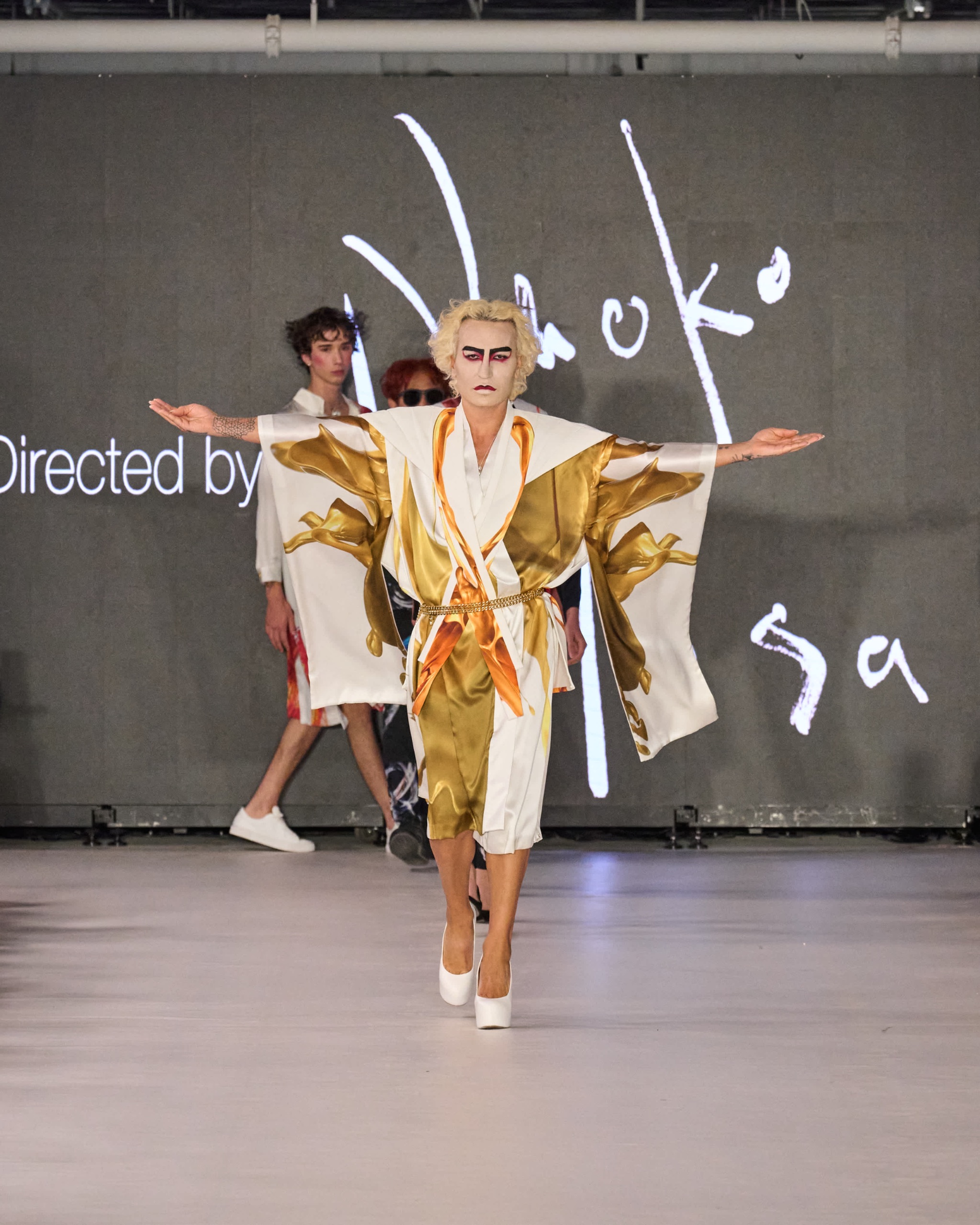 A model walking the runway wearing a gold and white kimono-inspired robe with florals patterns, belted at the waist.