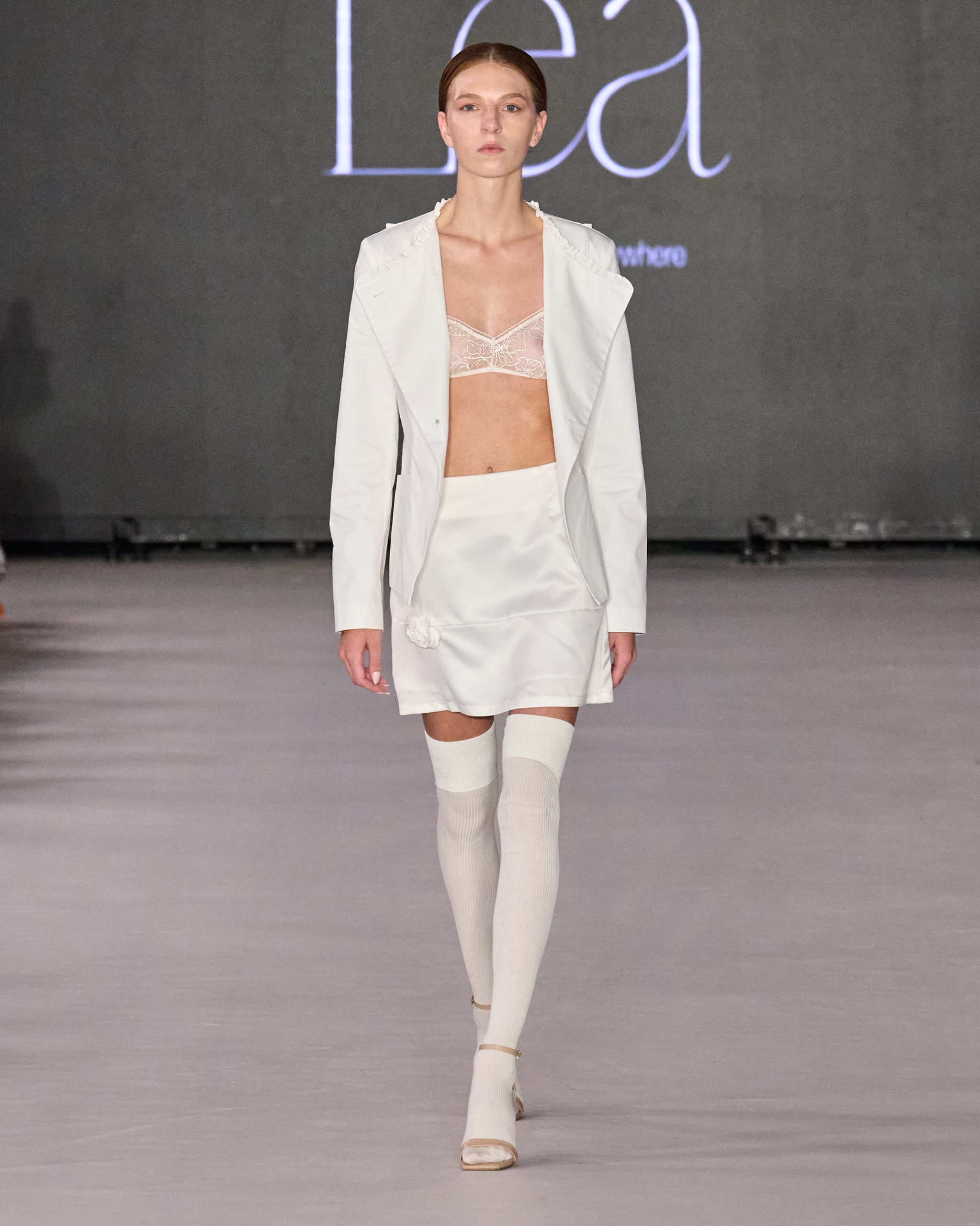 A model walking on the runway wearing a white bra, skirt, socks and jacket.