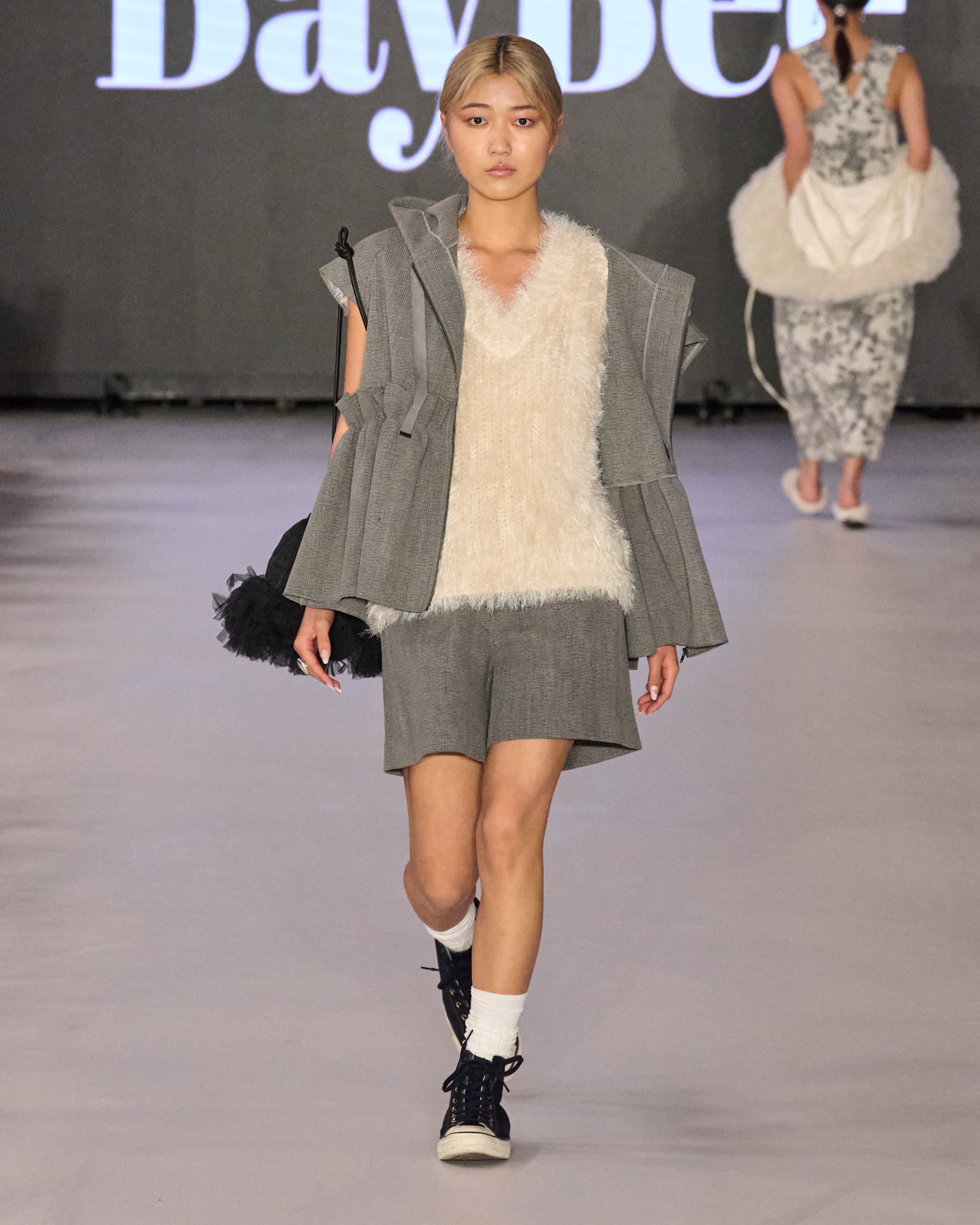 A model walking on the runway wearing a white jumper, grey shorts and jacket.