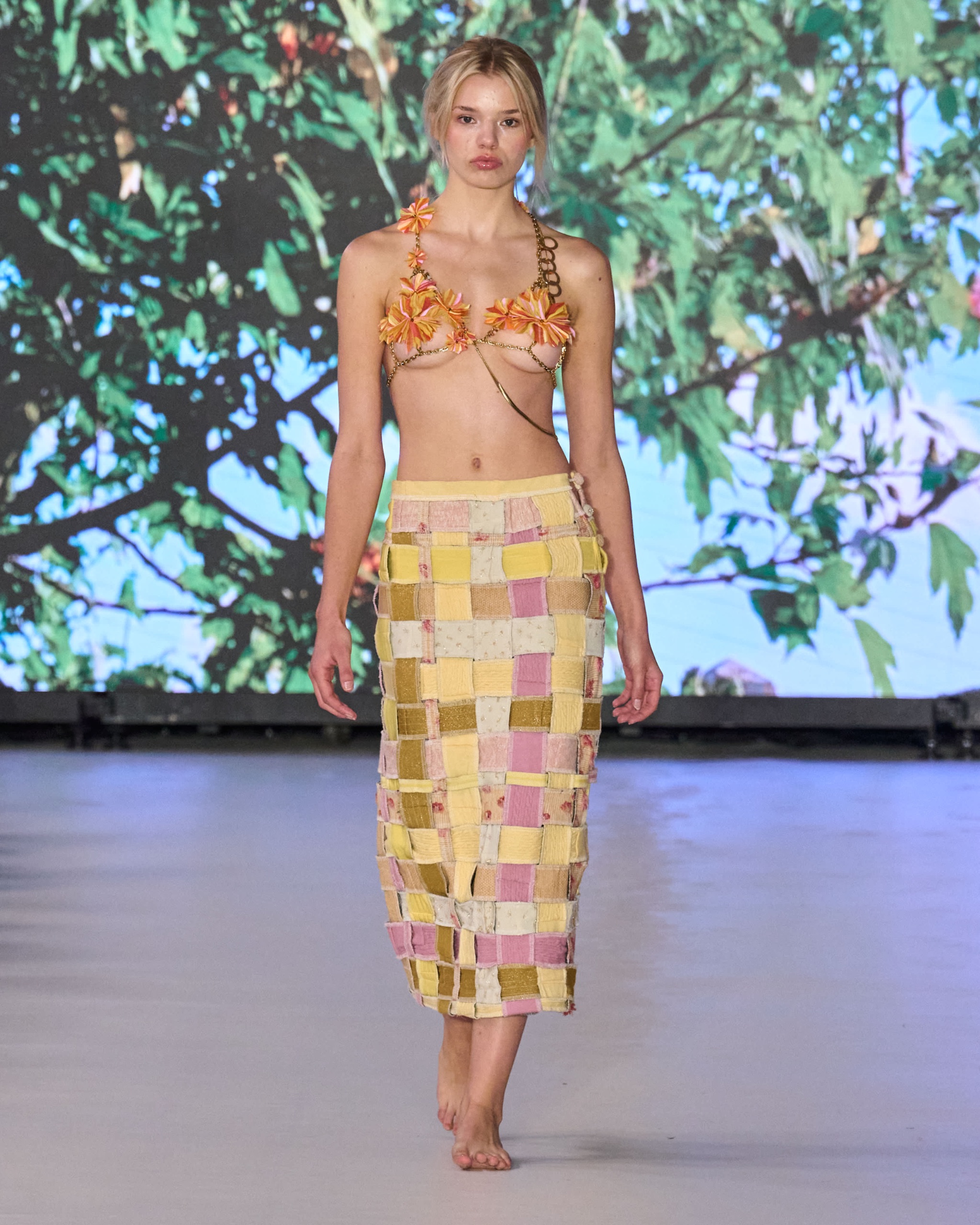 A model walking the runway wearing a orange bikini top with a yellow checkered skirt.