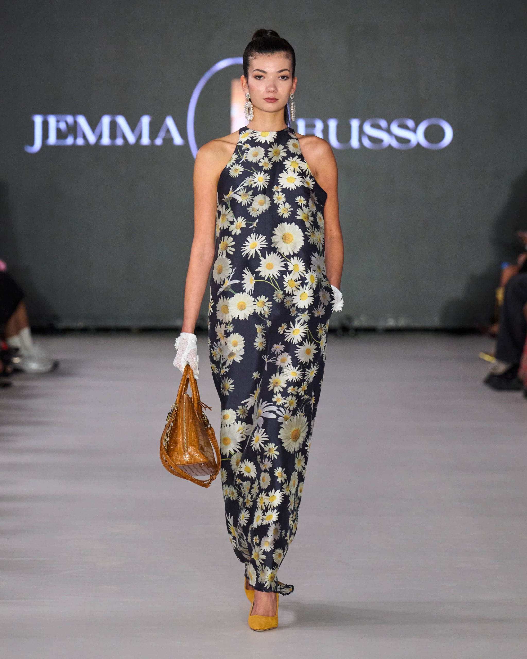 A model walking the runway wearing a floral jumpsuit with tan bag.