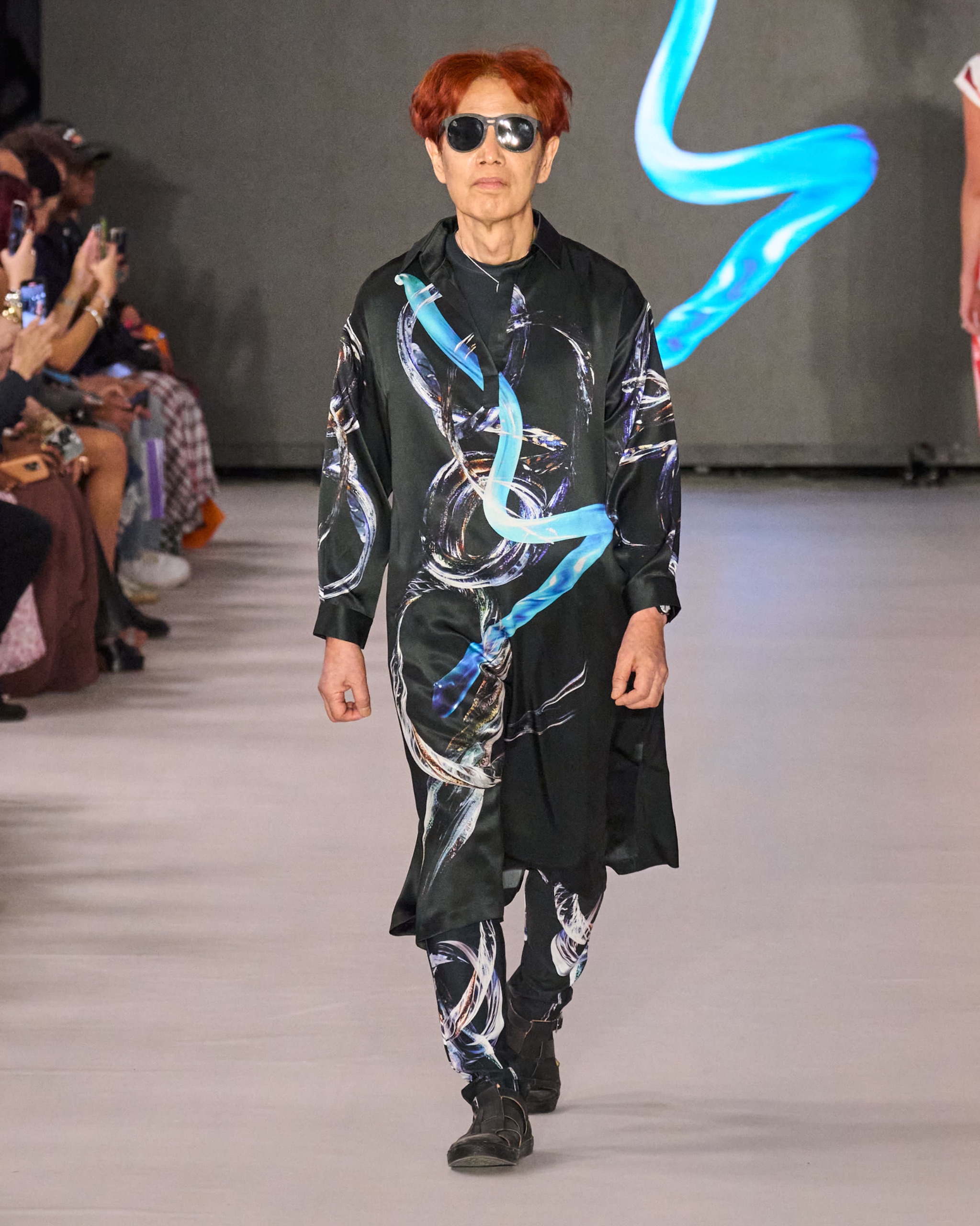 A model walking the runway wearing a black outfit with blue and grey detailing.