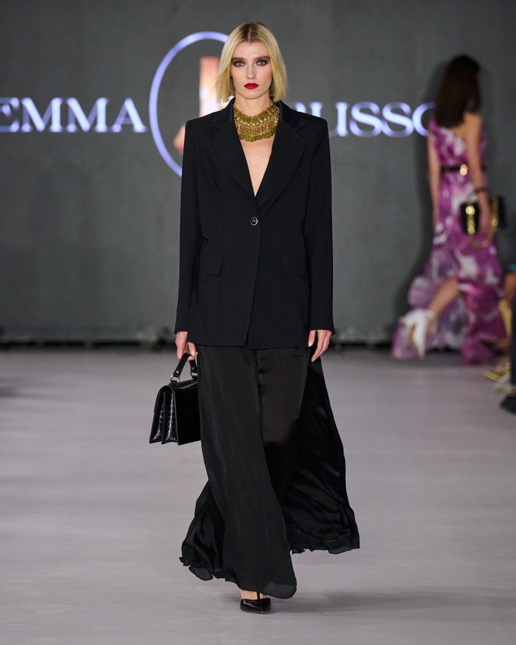 A model walking the runway wearing a black blazer, bag and trousers.