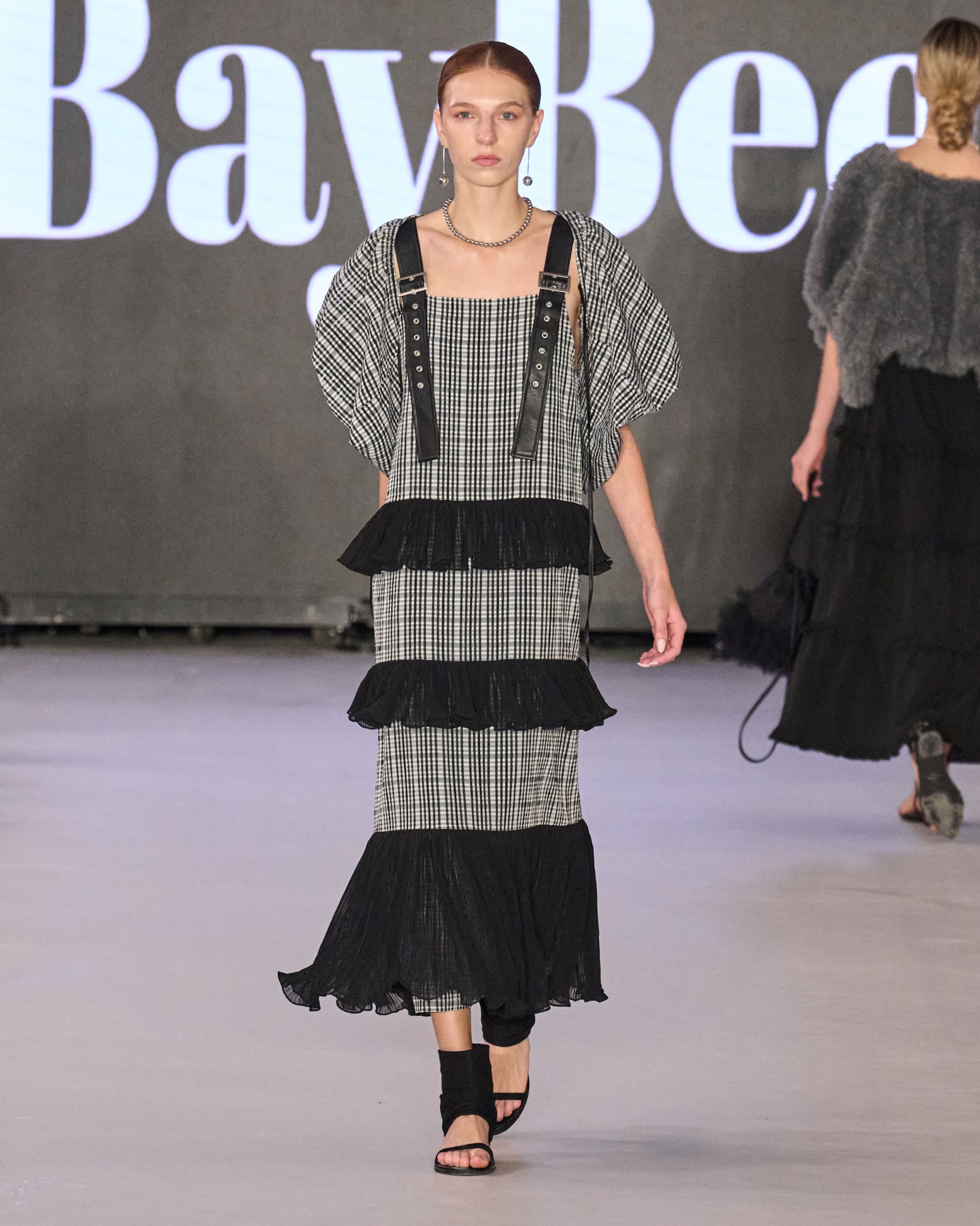 A model walking the runway wearing a checkered dress with puffed sleeves, black ruffles with black strap details.