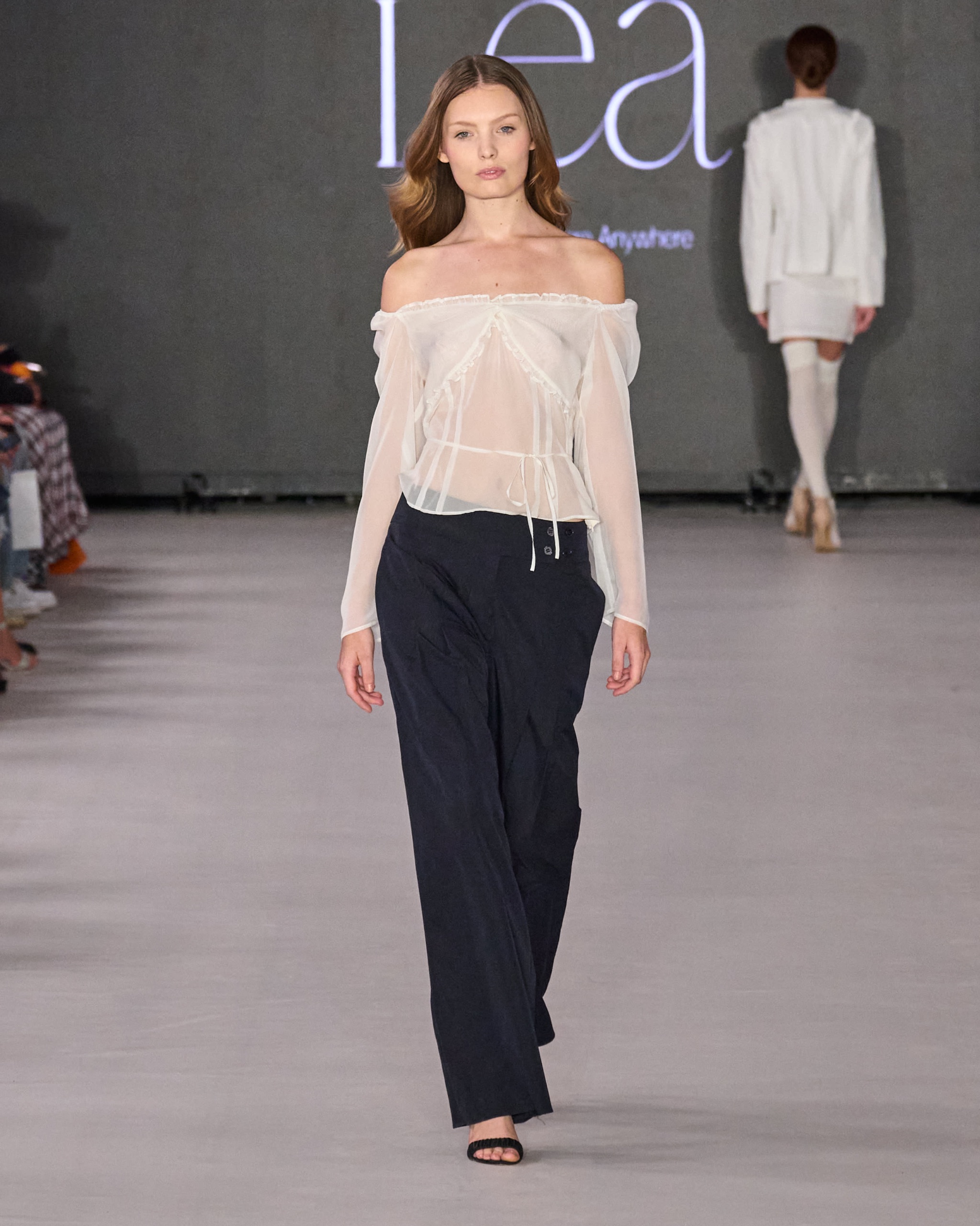 A model walking the runway wearing a sheer off-the-shoulder white top and black trousers.