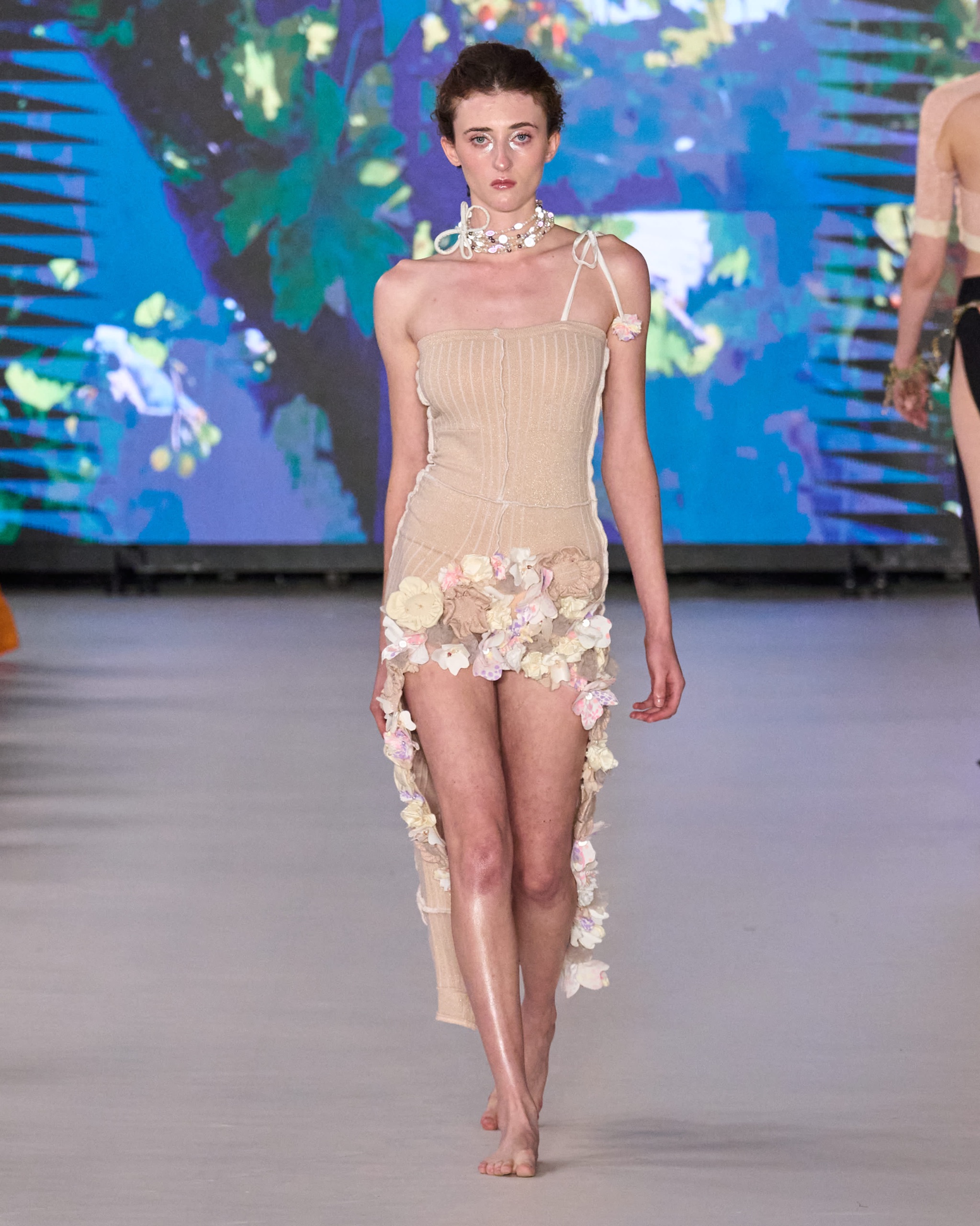 A model walking the runway wearing a tan dress with floral detailing.