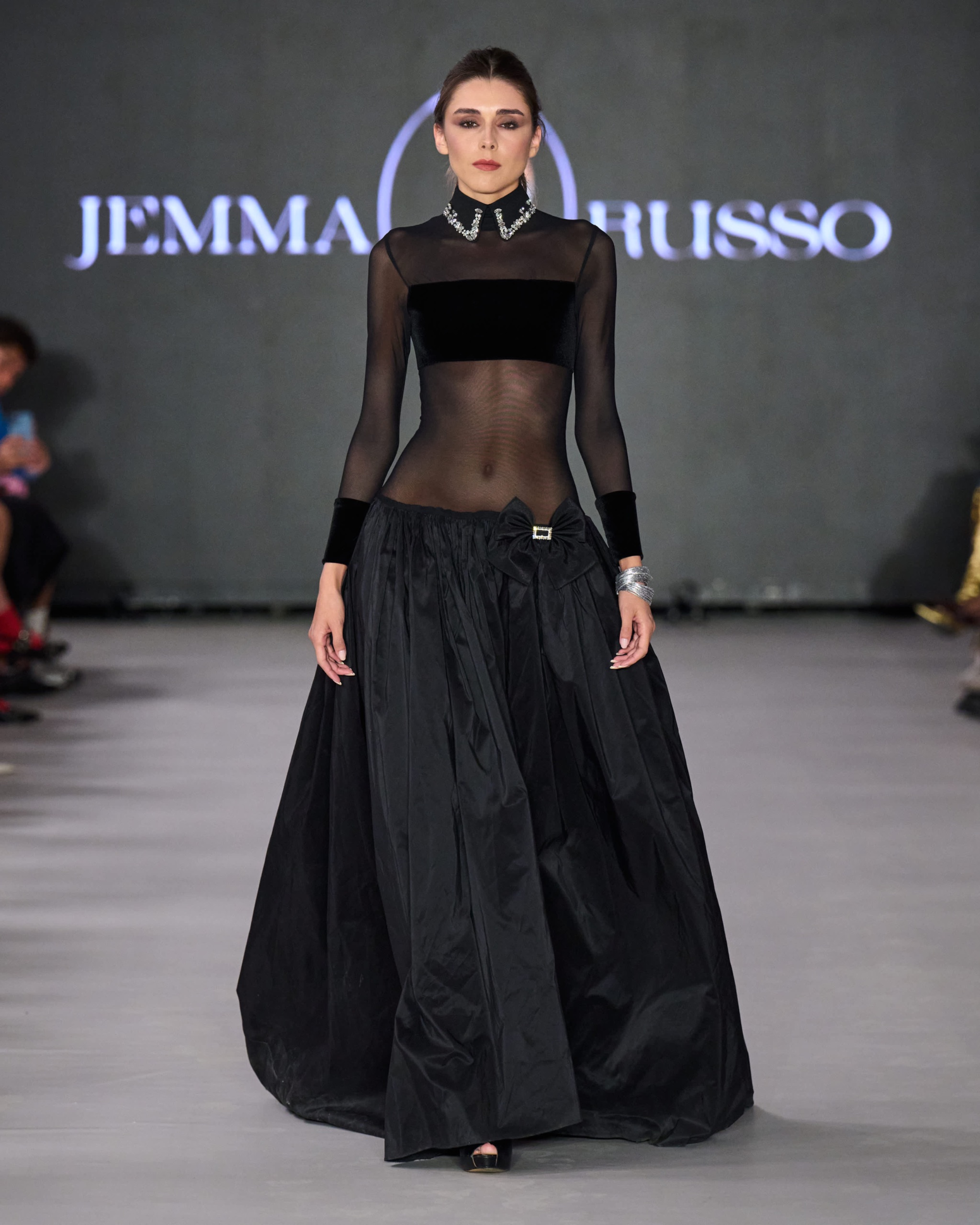 A model walking the runway wearing a long-sleeved black sheer top and long black skirt.