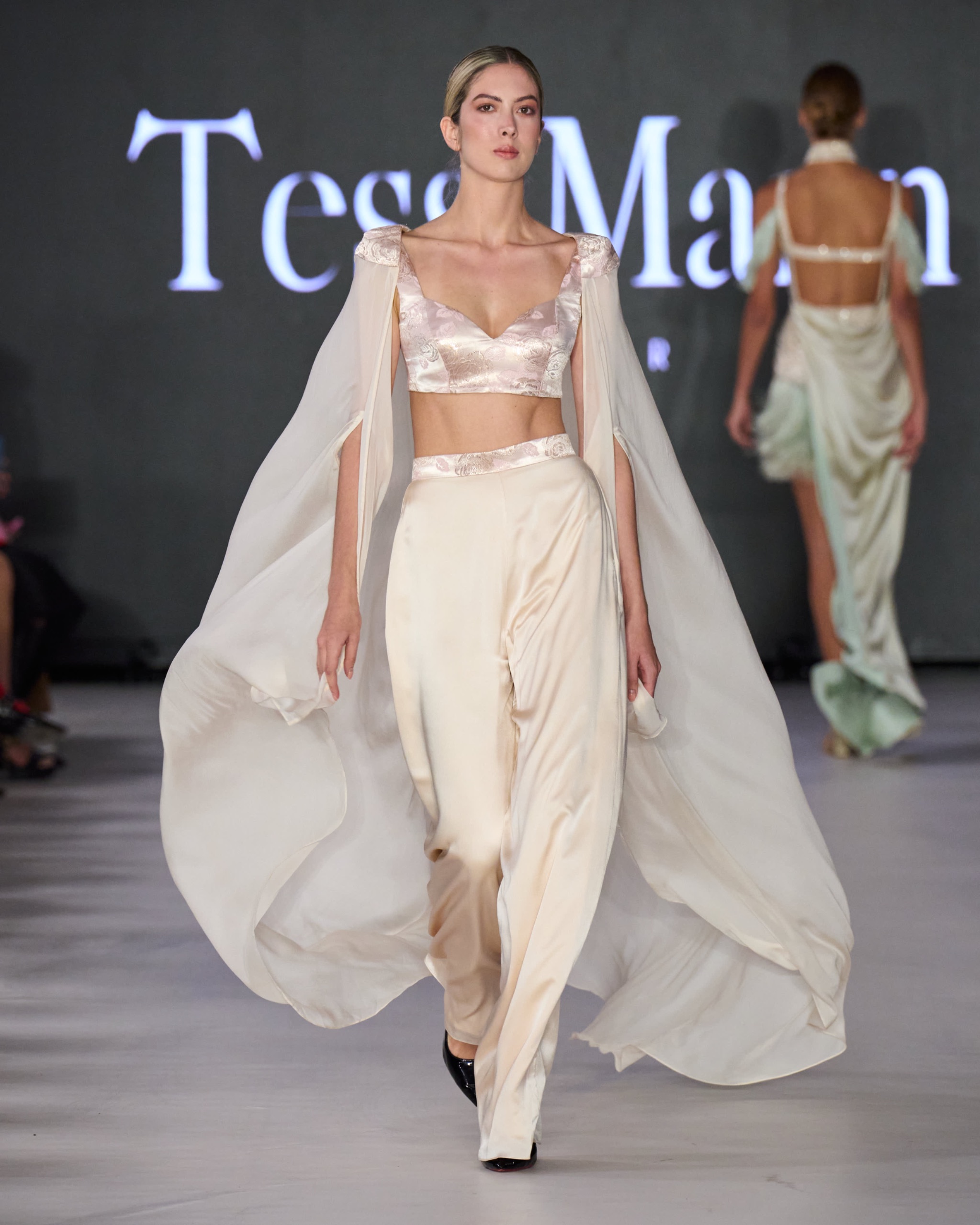 A model walking the runway wearing white trousers with a white top and a white flowy cape.