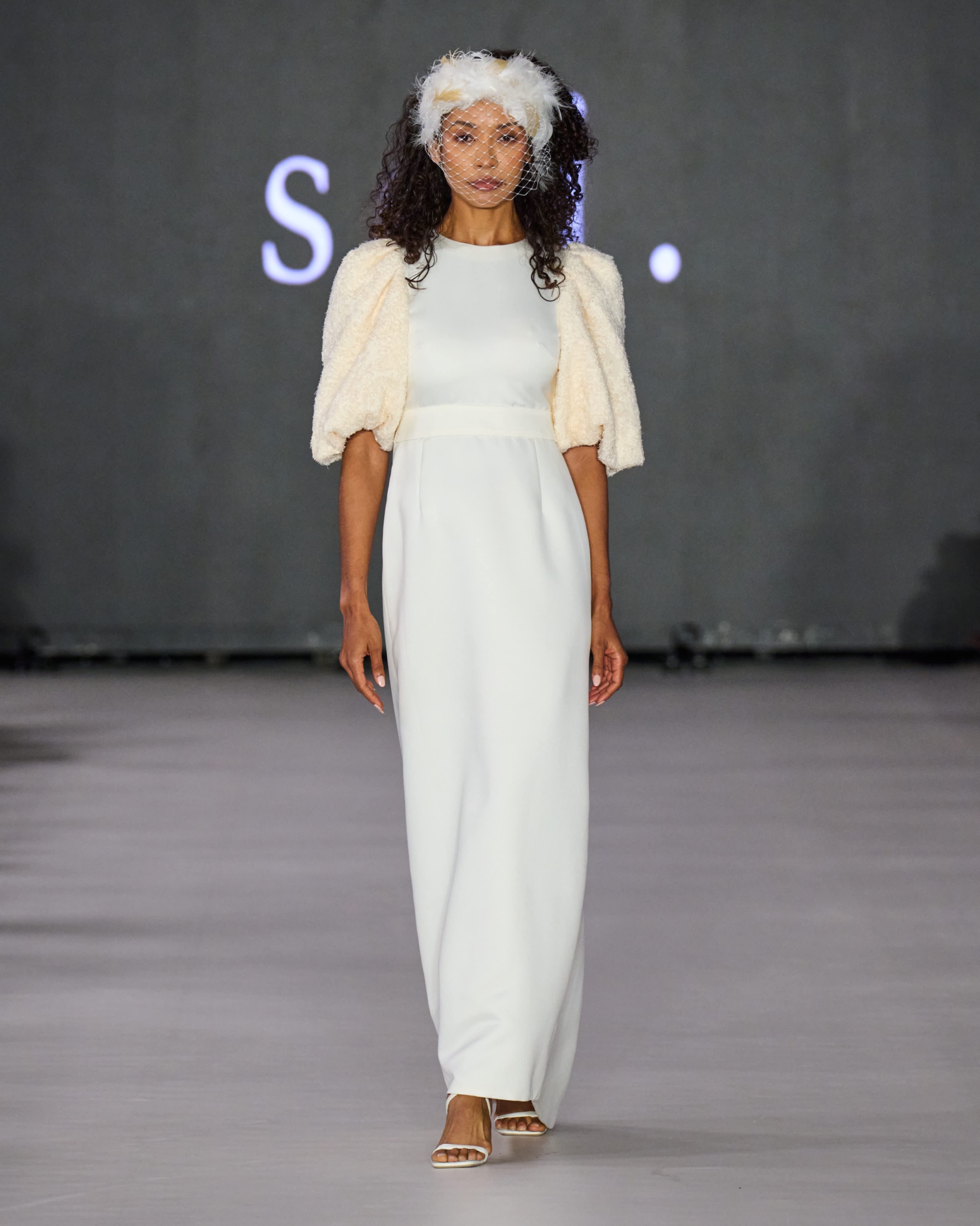 A model walking the runway wearing a white jumpsuit with cream jacket.