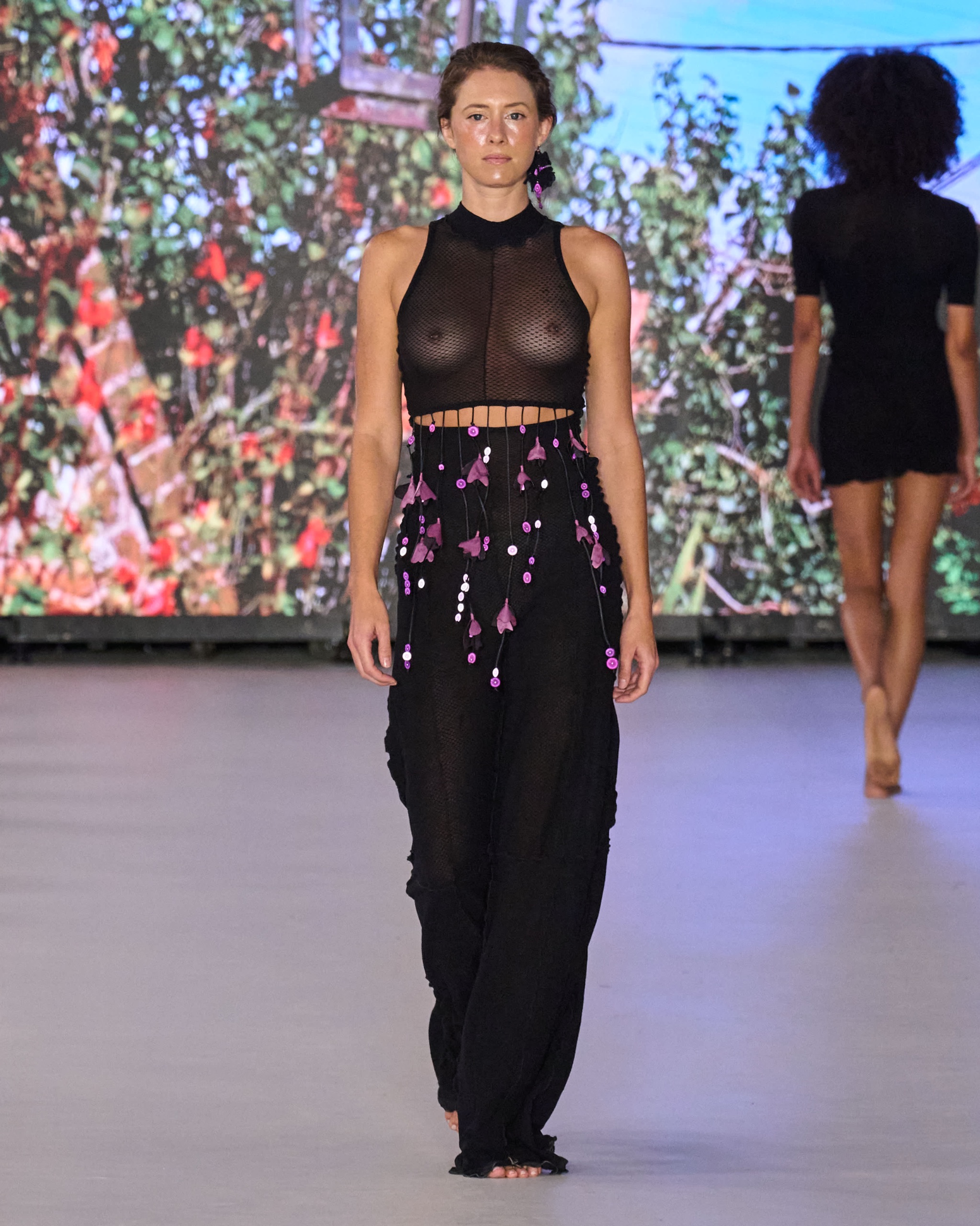 A model walking the runway wearing a black sheer top and black embellished trousers.