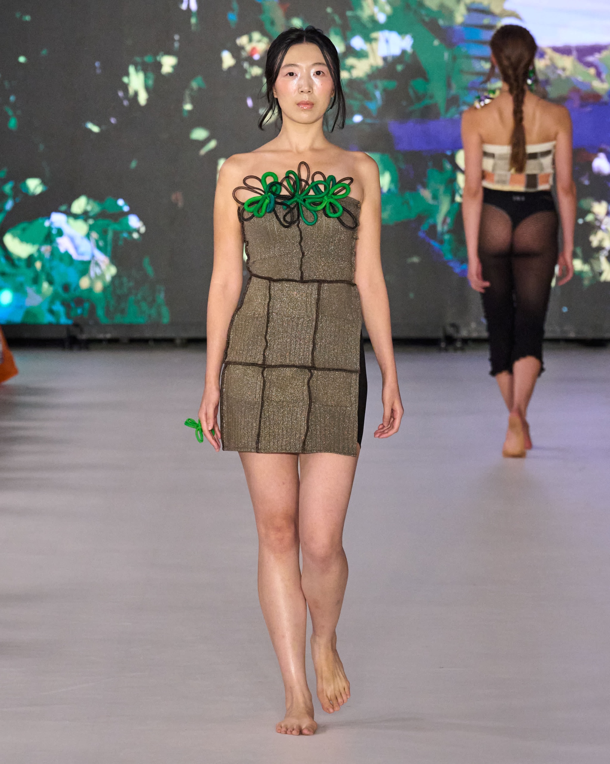 A model walking the runway wearing an olive grid patterned dress with green embellishments.