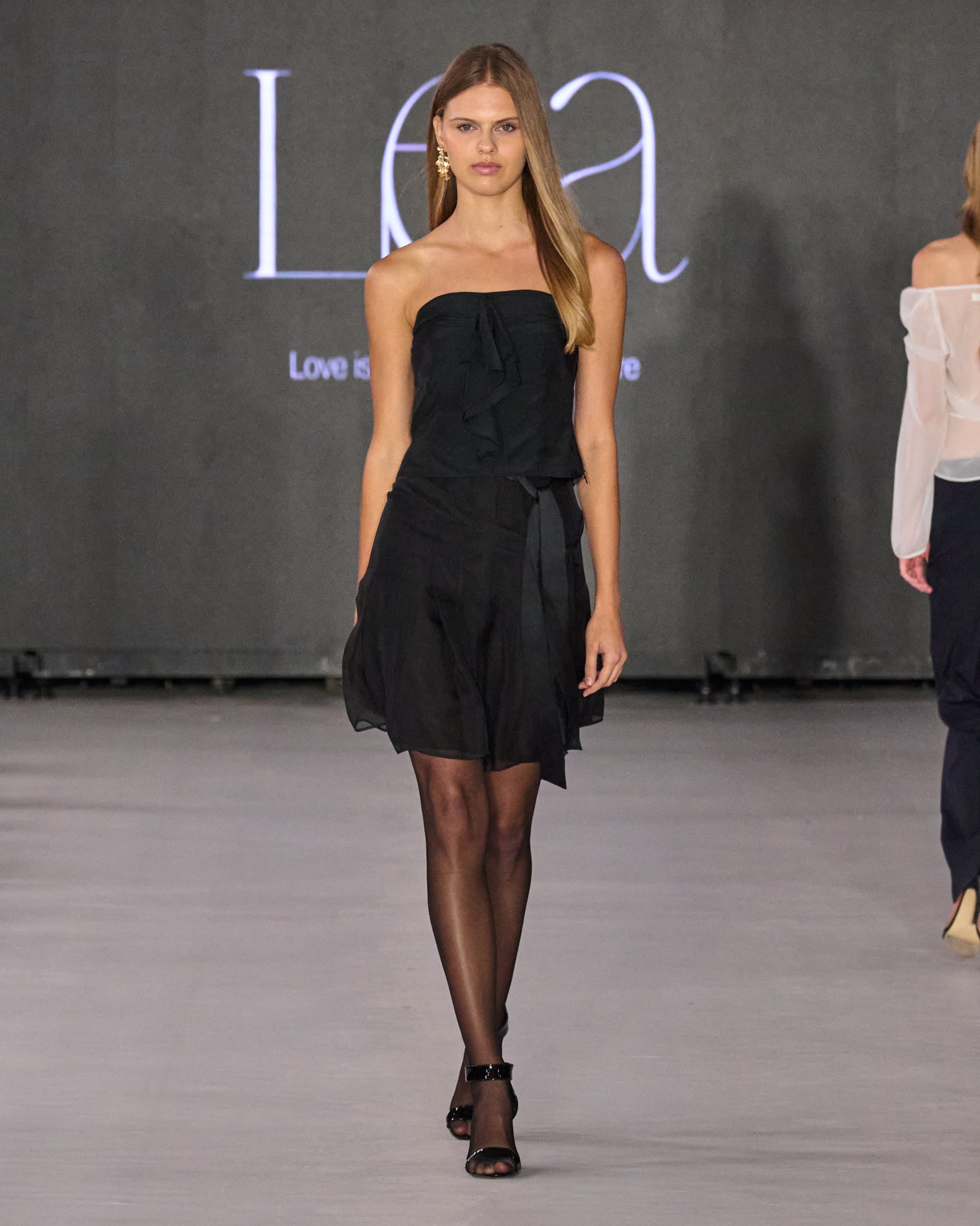 A model walking the runway wearing a black dress and tights.