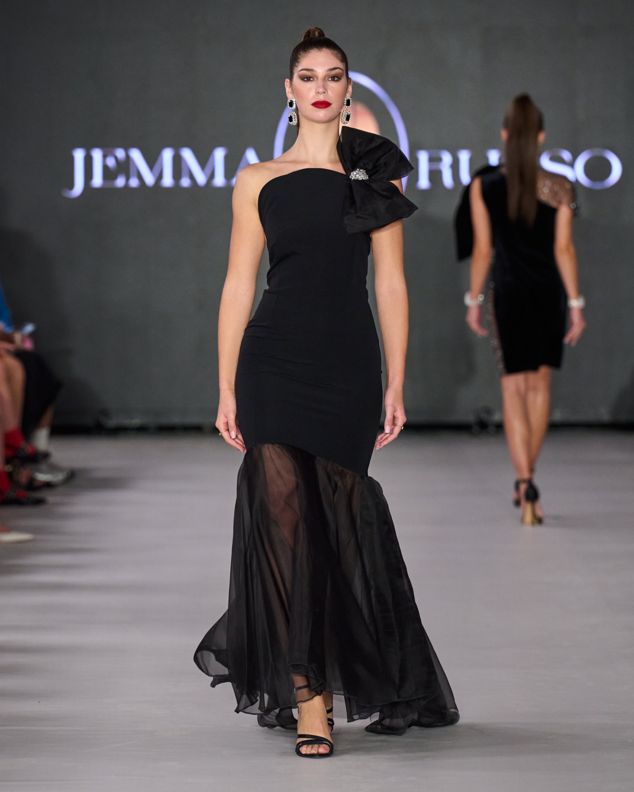 A model walking the runway wearing a long black dress with bow detail.