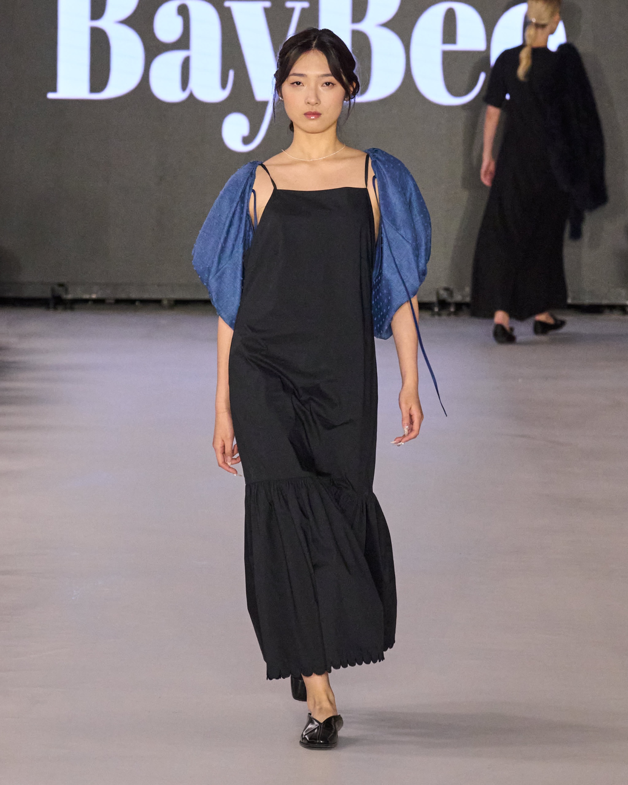 A model walking the runway wearing a long black dress with blue jacket.