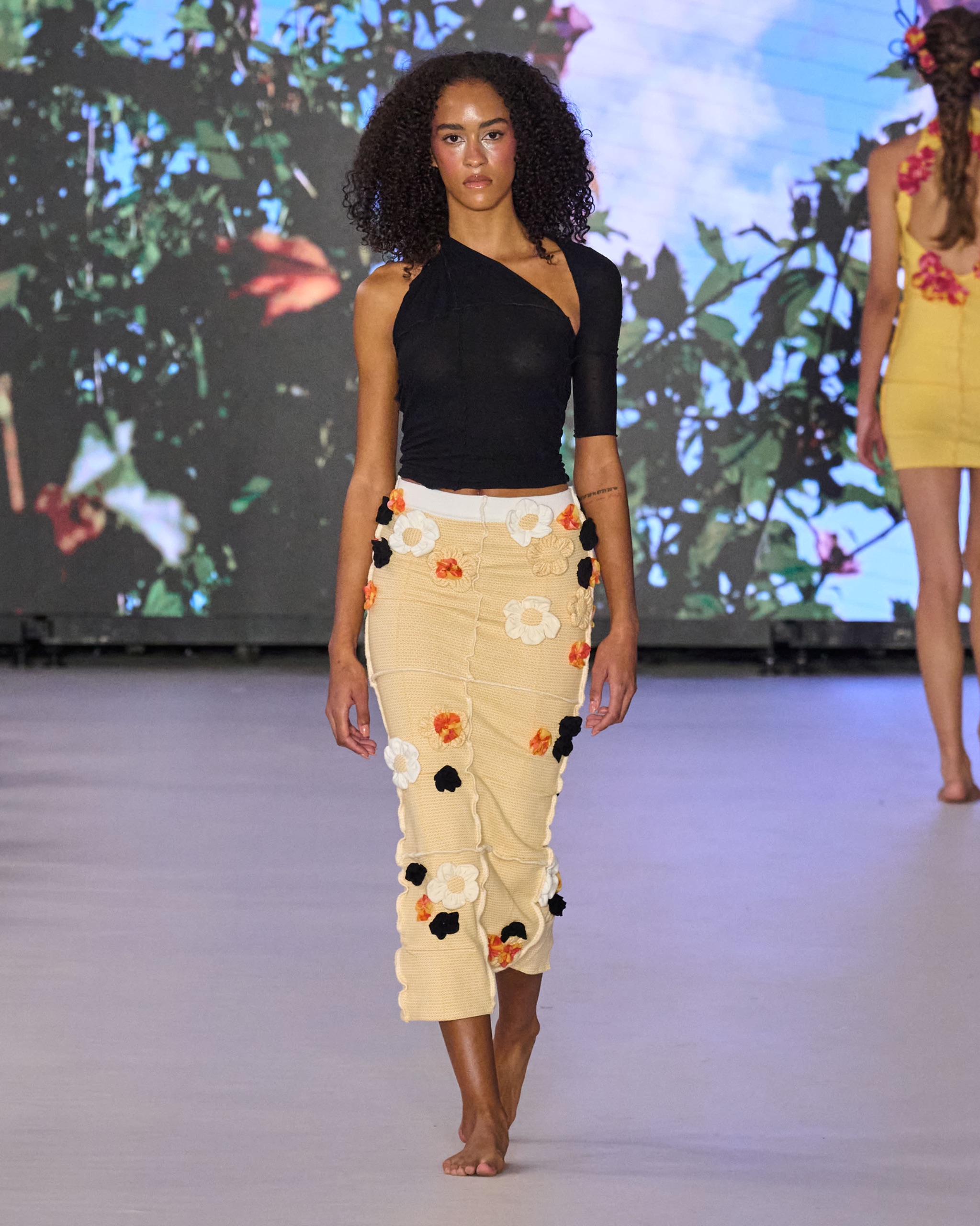 A model walking the runway wearing a one sleeved black top and yellow skirt with orange, white and black detailing.