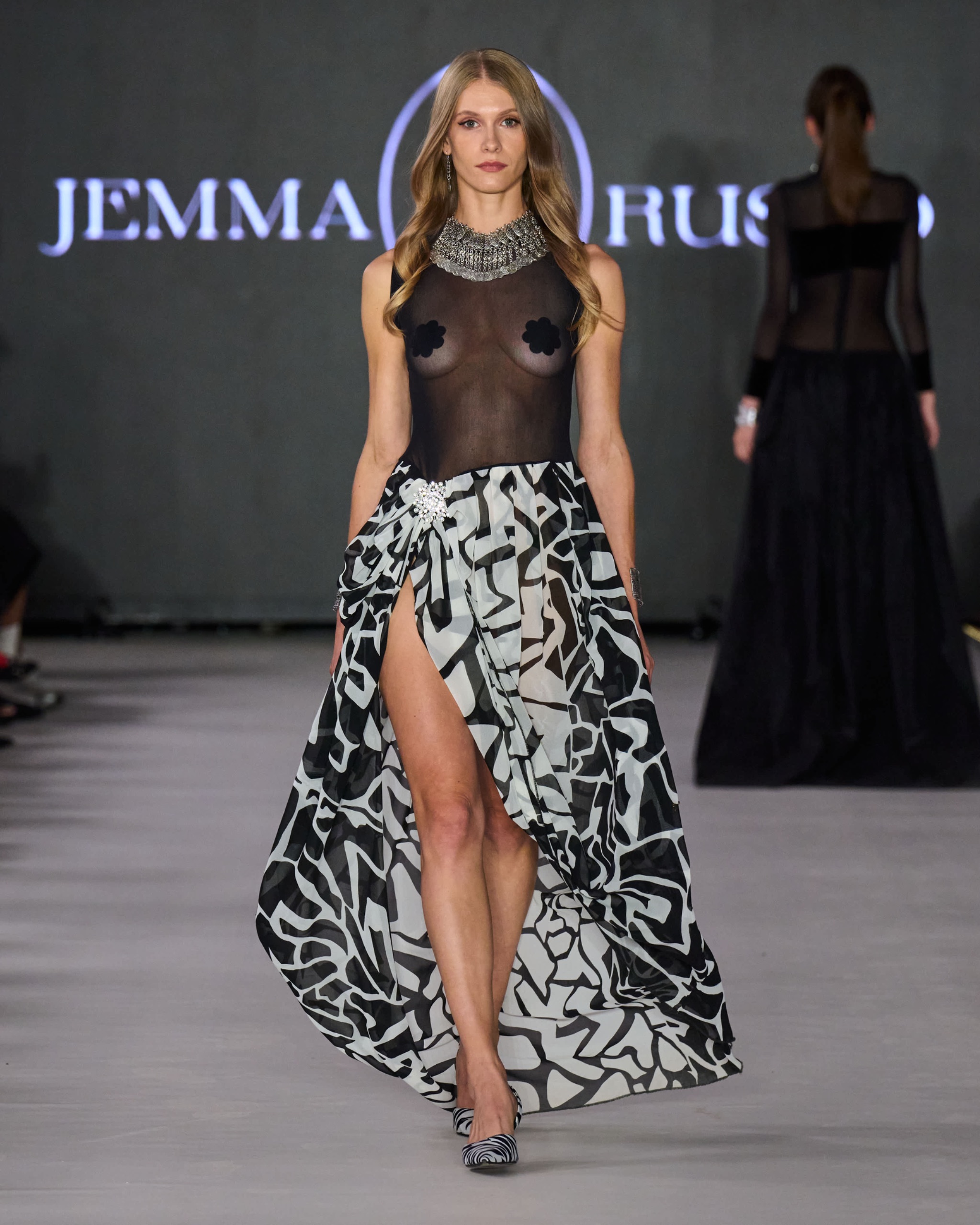 A model walking the runway wearing a high neck sheer top with a black and white swirl patterned skirt.