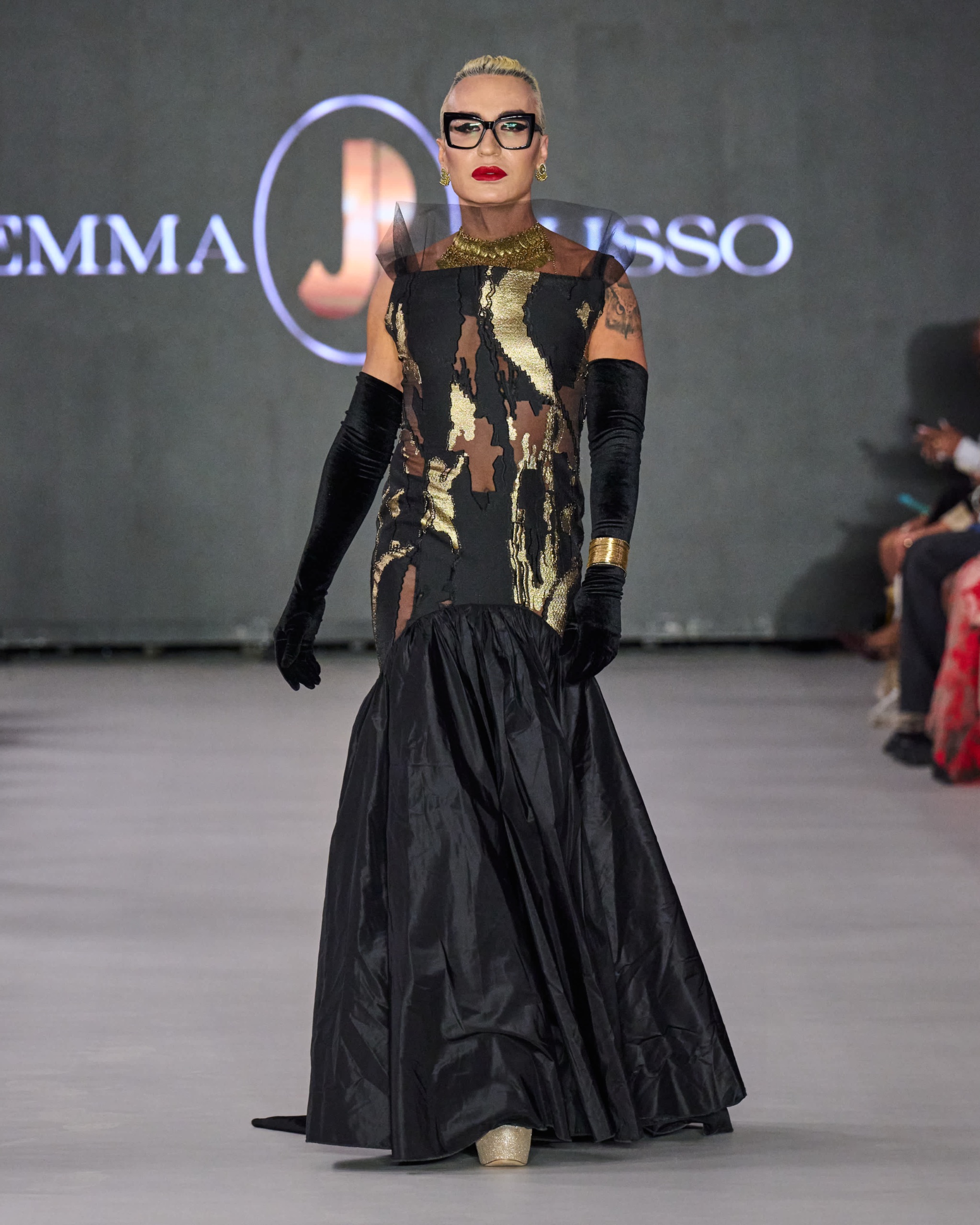 A model walking the runway wearing a long black dress with gold detailing and long black gloves.