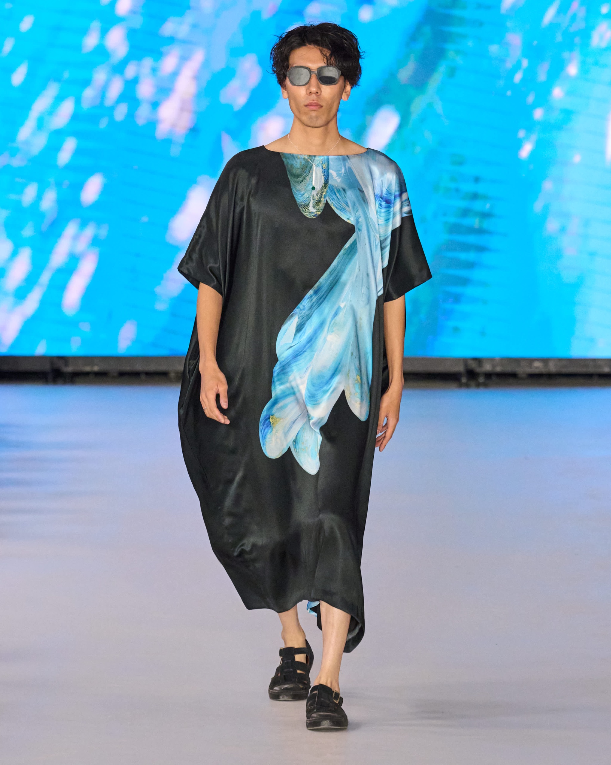 A model walking the runway wearing a long black dress with blue and white detailing.